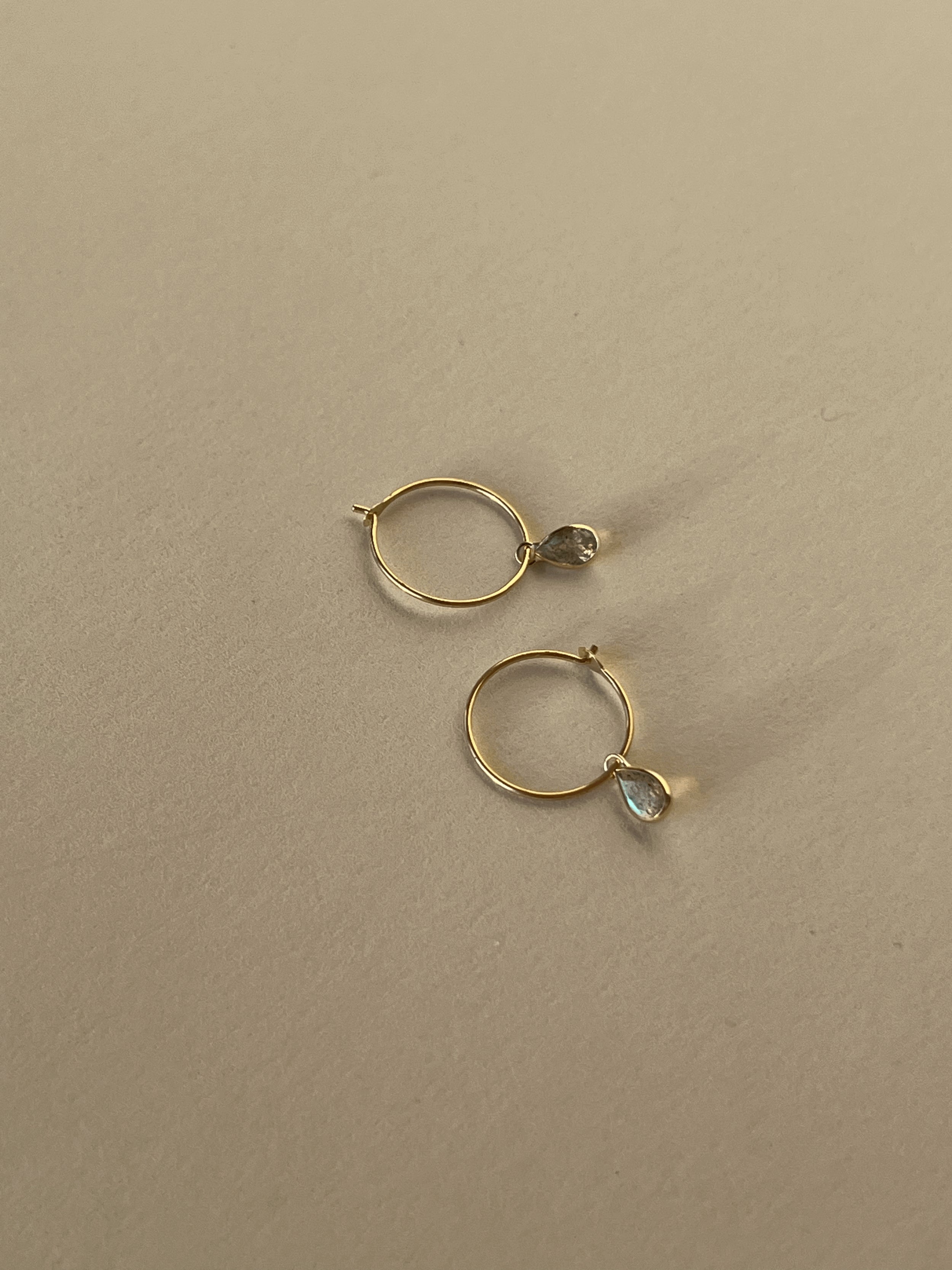 Product shot of Thalken 14k gold 10mm hoop earrings paired with small pear-shaped drop charms, showcasing minimalist and timeless design