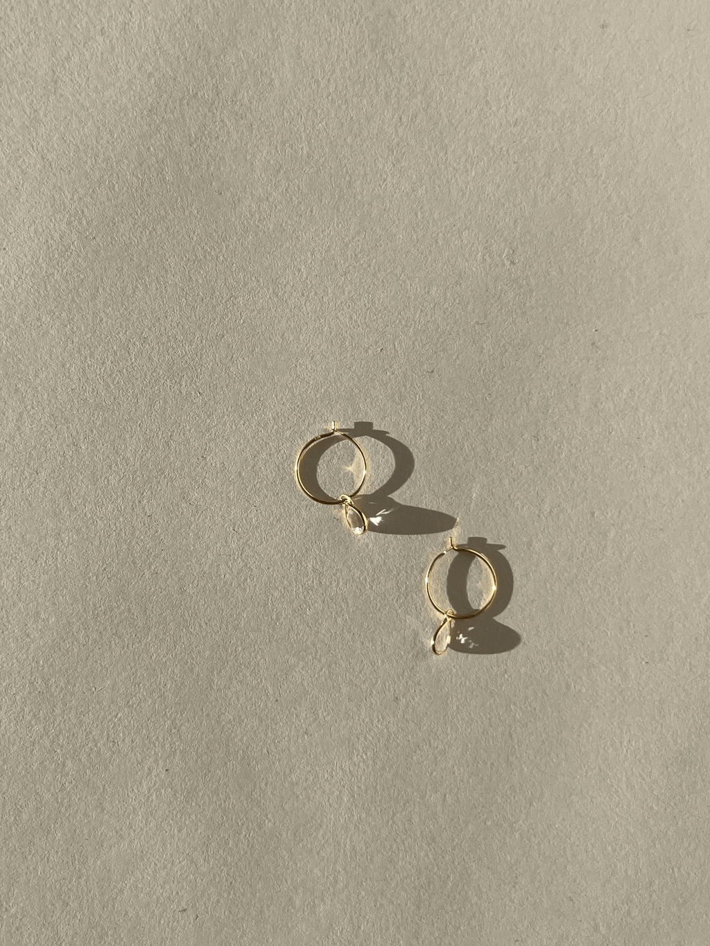 Product shot of Thalken 14k gold 10mm hoop earrings with small pear-shaped white topaz drop charms, highlighting minimalist fine jewelry design