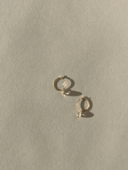 Product shot of Thalken 14k gold 10mm hoop earrings with small pear-shaped white topaz drop charms, highlighting minimalist fine jewelry design