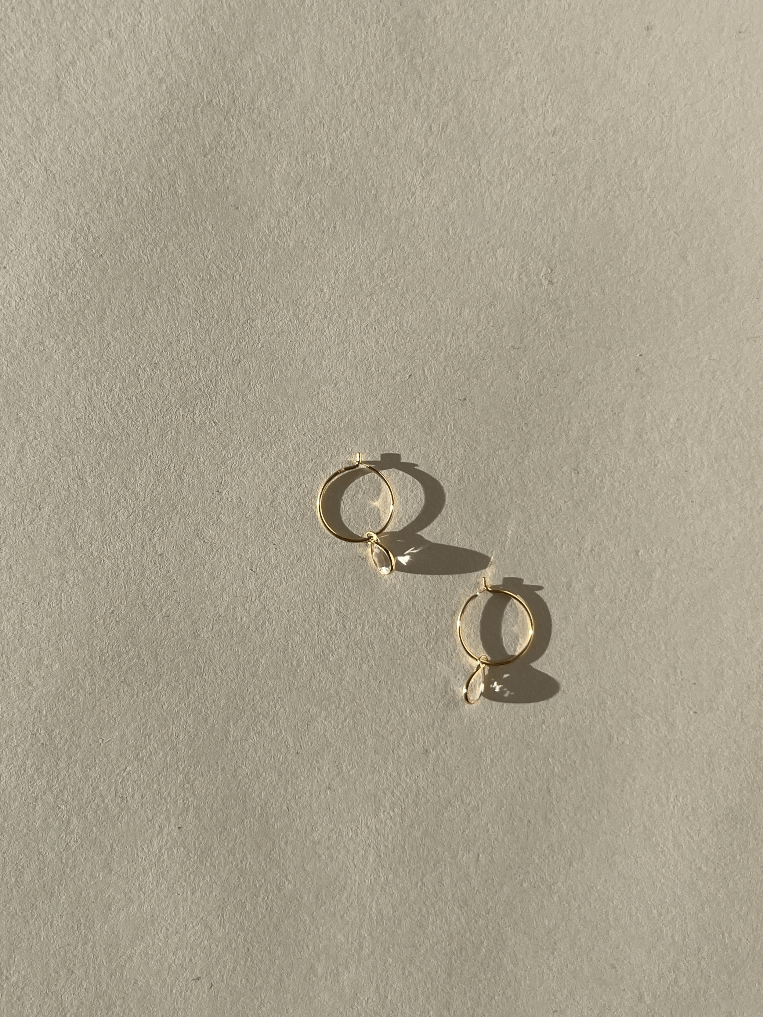 Product shot of Thalken 14k gold 10mm hoop earrings with small pear-shaped white topaz drop charms, highlighting minimalist fine jewelry design
