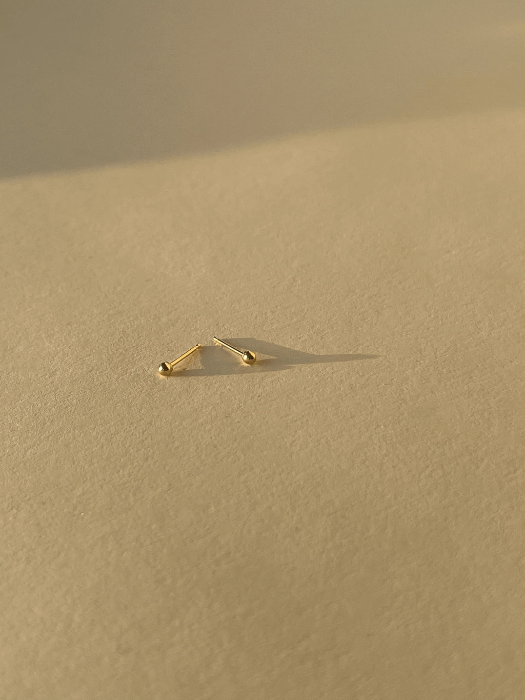 Close-up of 2mm 14k solid gold ball stud earring pair on a neutral background, highlighting their petite size and polished finish
