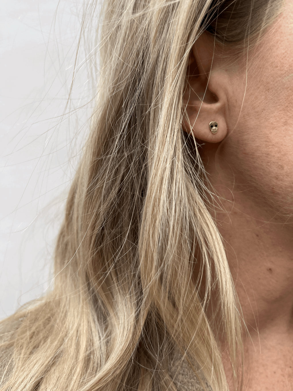 Close-up of model wearing a single Tiny Skull Stud Earring in her first ear piercing; bold black enamel eyes visible on minimalist gold skull design