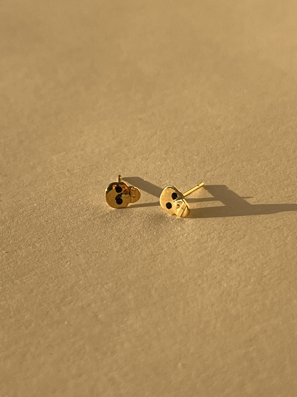 Close-up of a pair of Tiny Skull Stud Earrings displayed on a neutral background, showcasing gold skull shape and black enamel eye detailing