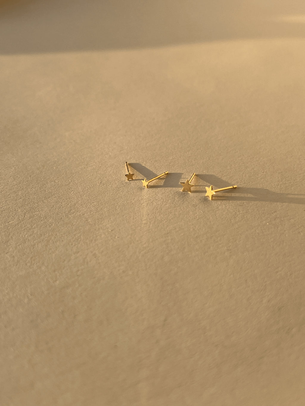 Wide shot of the 3mm and 4mm Tiny Star Stud Earrings in 14k solid yellow gold placed together on a soft, neutral surface