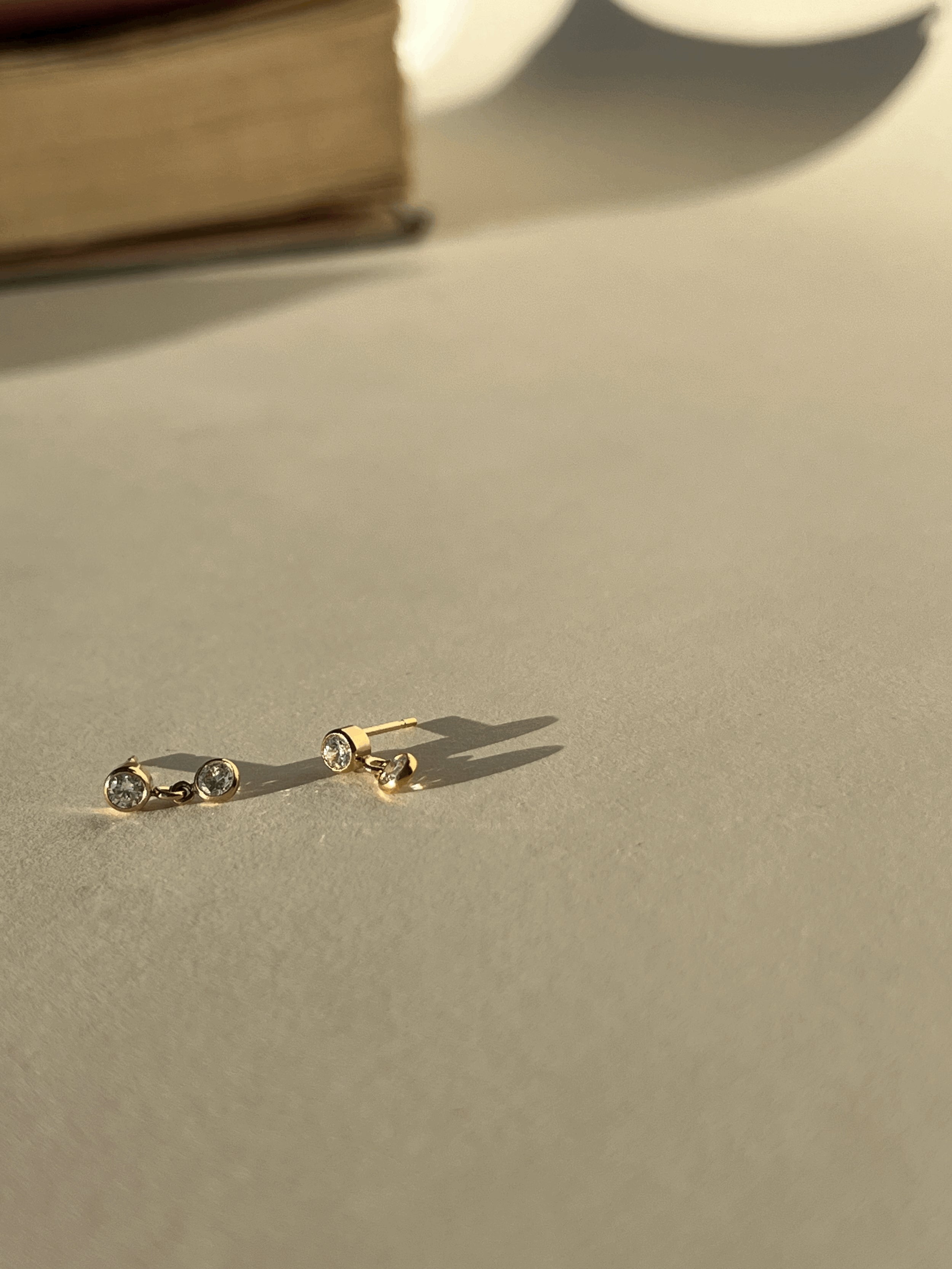  Artful product photo of Thalken’s gold-filled cubic zirconia stud and drop earrings with a book in the background, creating a warm lifestyle aesthetic