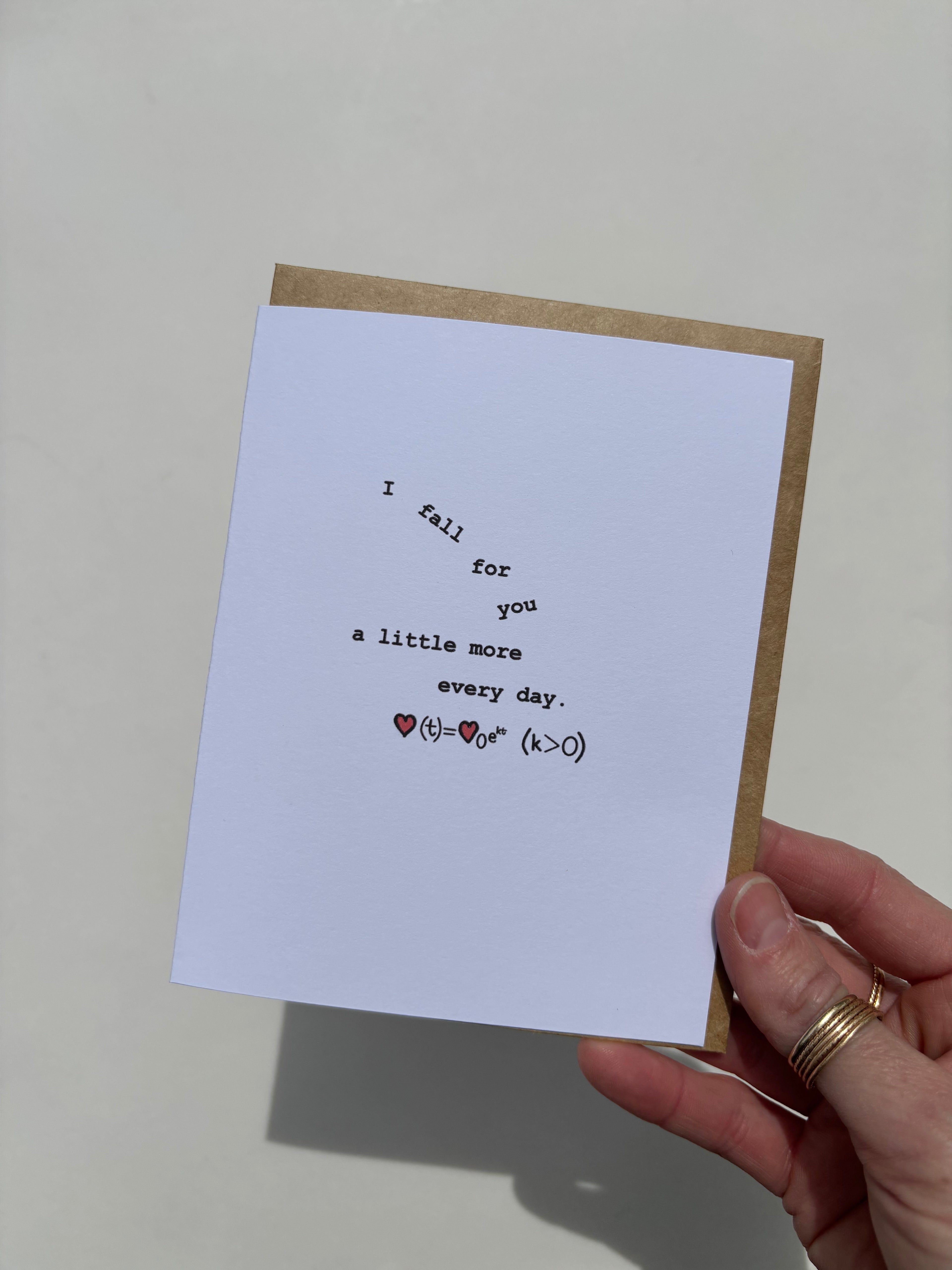 Greeting card held in hand reading “I fall for you a little more every day,” featuring a symbolic equation inspired by the continuous growth model, with the words “I fall for you” arranged to appear as if they’re gently falling.