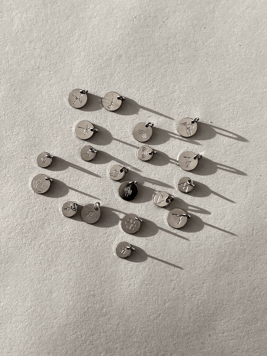 Black and white image of sterling silver disc charms stamped with designs including dandelion seed, mountains, dog paw, rainbow, tree, compass, cactus, Hawaiian Islands, and lotus flower, photographed in both 7mm and 9mm sizes except for the Mountains with Sun, Compass, and Hawaiian Islands designs, which are shown in 9mm only
