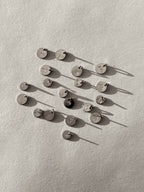 Black and white image of sterling silver disc charms stamped with designs including dandelion seed, mountains, dog paw, rainbow, tree, compass, cactus, Hawaiian Islands, and lotus flower, photographed in both 7mm and 9mm sizes except for the Mountains with Sun, Compass, and Hawaiian Islands designs, which are shown in 9mm only