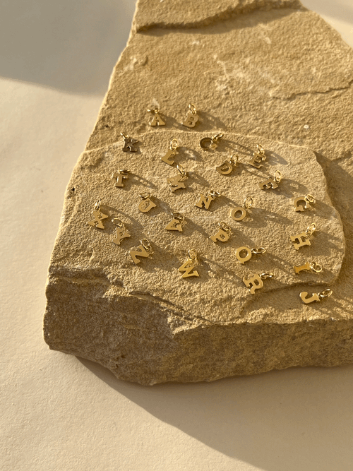 Tiny gold-filled block letter charms displayed on a natural rock surface