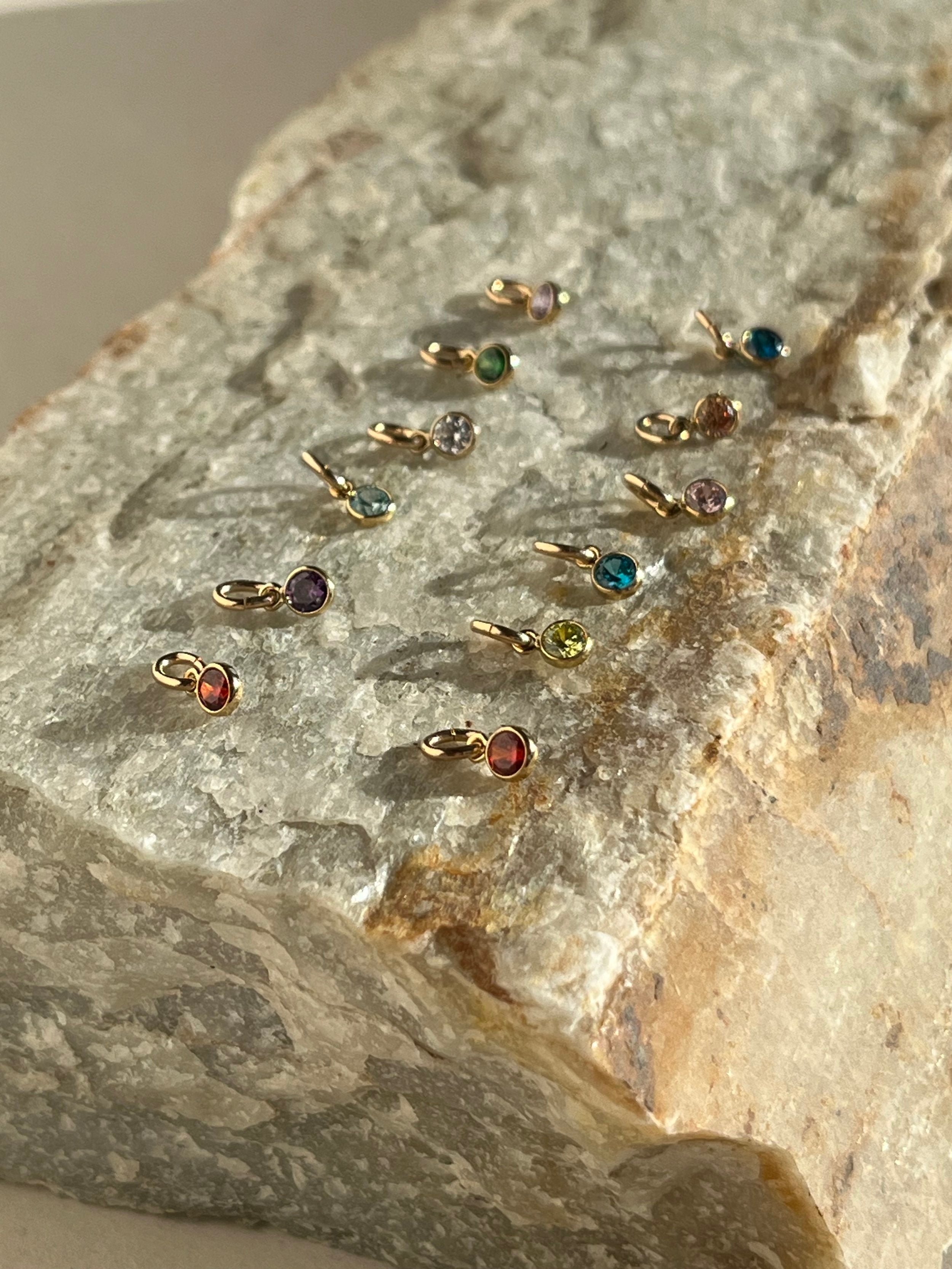 A slightly pulled-back view of twelve yellow gold-filled 3mm cubic zirconia birthstone charms in bezel settings, arranged on a natural rock