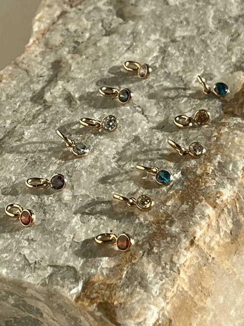 Twelve 3mm yellow gold-filled cubic zirconia birthstone charms in bezel settings, displayed in a close-up on a textured rock surface