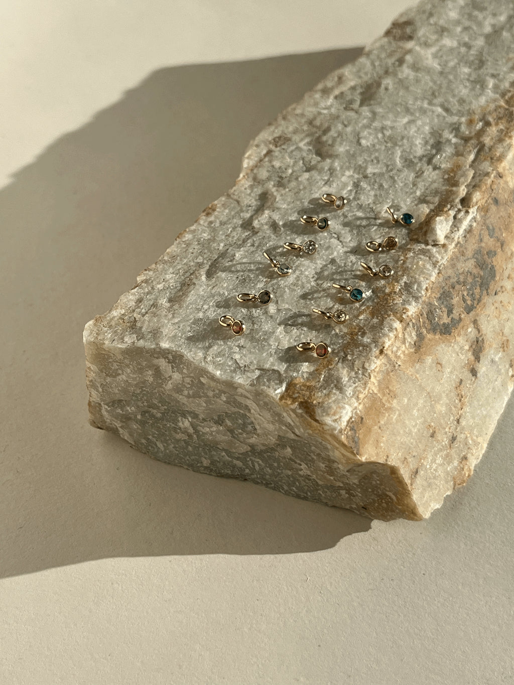 Twelve yellow gold-filled 3mm bezel-set cubic zirconia birthstone charms displayed on a rock, photographed from a wider angle to show color variety