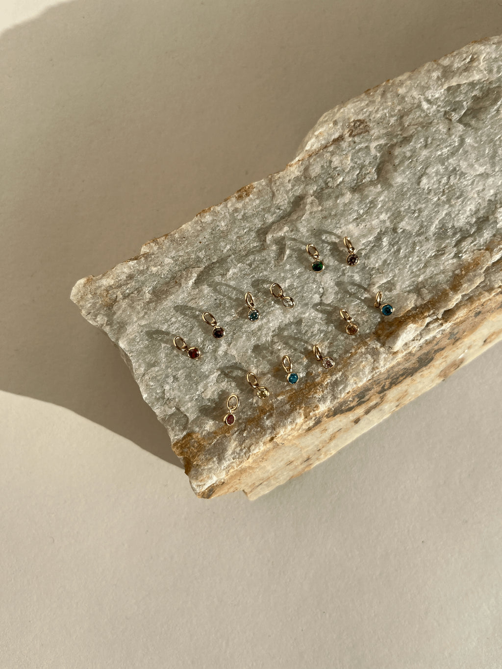 A different angled view of twelve 3mm yellow gold-filled cubic zirconia birthstone charms in bezel settings, arranged on a rock surface