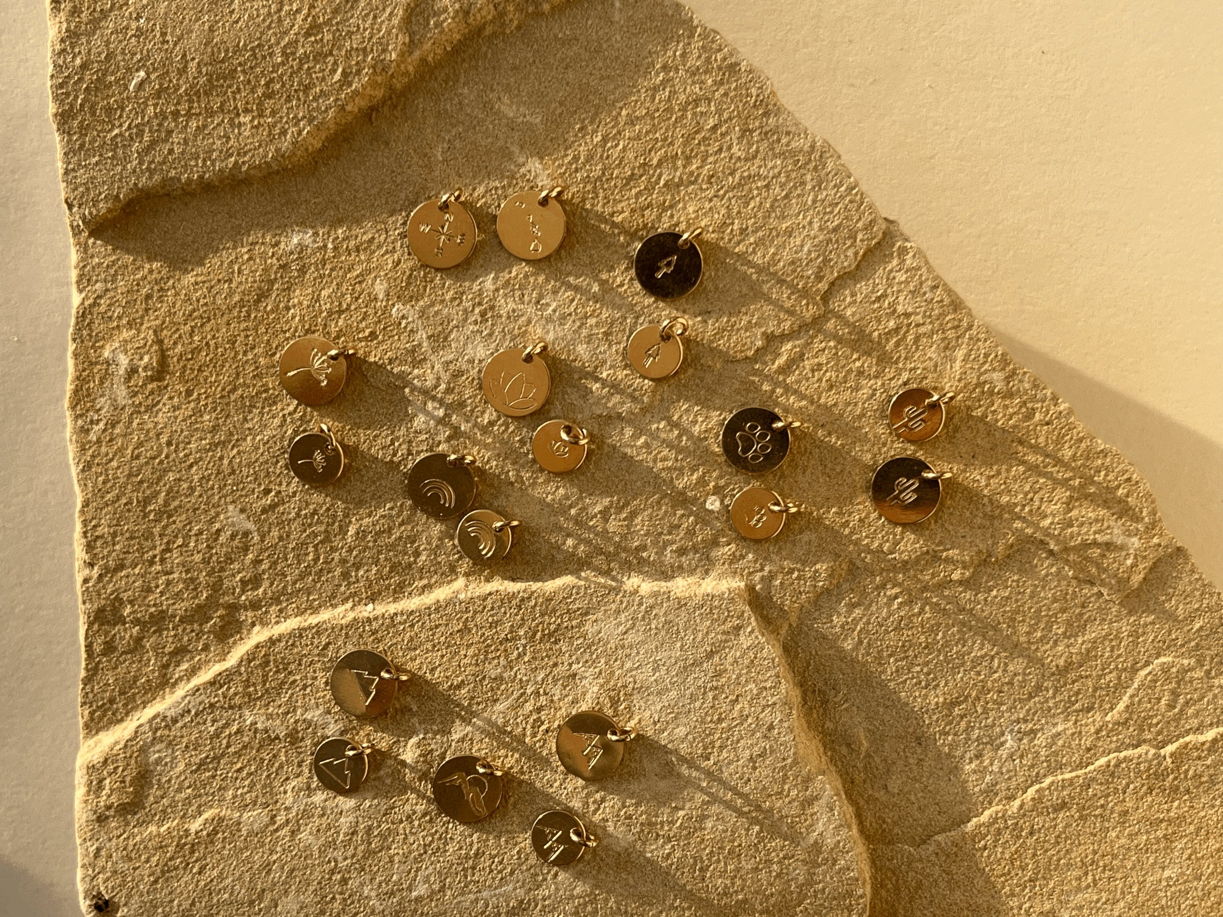 Gold-filled disc charms displayed on a rock, featuring tree, dandelion, lotus, paw print, rainbow, three-peak mountains, and two-peak mountains designs in 7mm and 9mm sizes, plus Hawaiian Islands, compass, and mountains with sun designs in 9mm only
