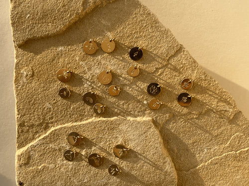 Gold-filled disc charms displayed on a rock, featuring tree, dandelion, lotus, paw print, rainbow, three-peak mountains, and two-peak mountains designs in 7mm and 9mm sizes, plus Hawaiian Islands, compass, and mountains with sun designs in 9mm only
