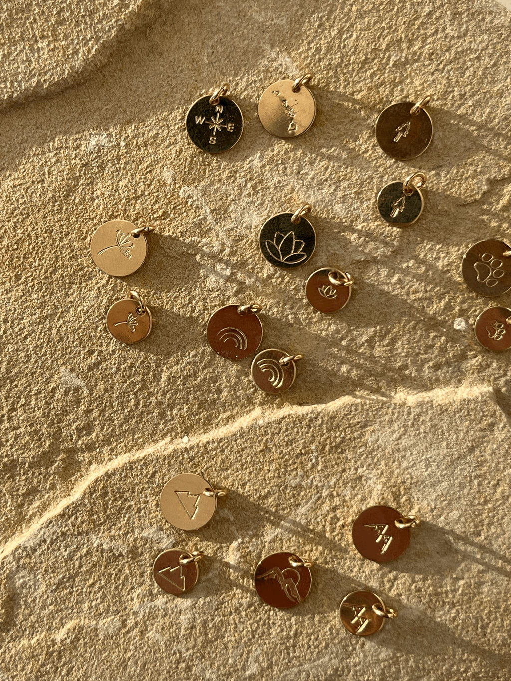 Gold-filled disc charms displayed on a rock, featuring tree, dandelion, lotus, paw print, rainbow, three-peak mountains, and two-peak mountains designs in 7mm and 9mm sizes, plus Hawaiian Islands, compass, and mountains with sun designs in 9mm only