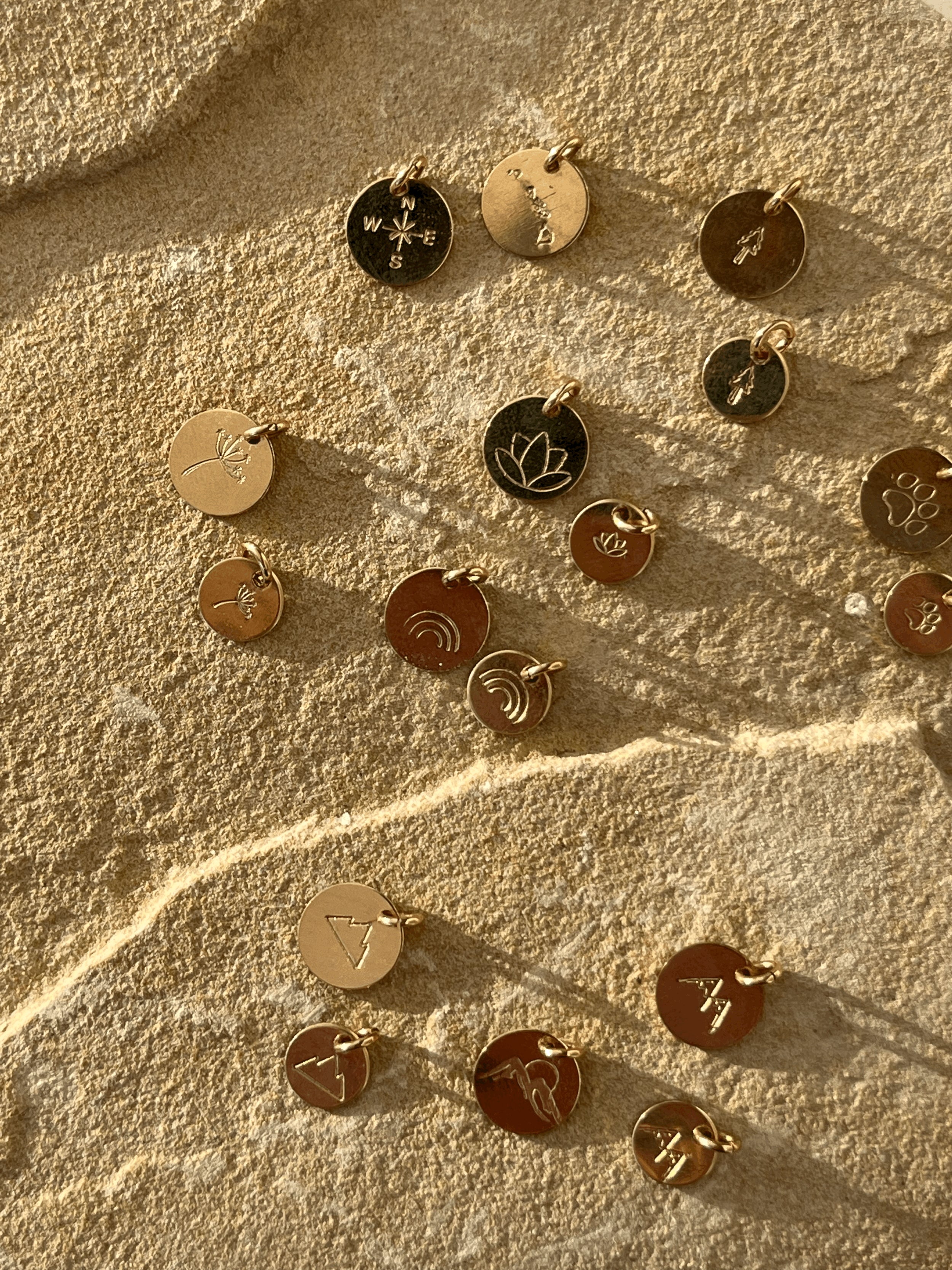 Gold-filled disc charms displayed on a rock, featuring tree, dandelion, lotus, paw print, rainbow, three-peak mountains, and two-peak mountains designs in 7mm and 9mm sizes, plus Hawaiian Islands, compass, and mountains with sun designs in 9mm only