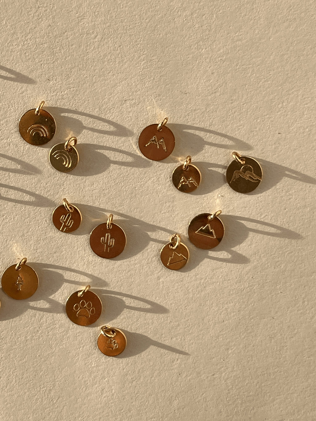 Gold-filled disc charms displayed on a rock, featuring tree, paw print, rainbow, cactus, three-peak mountains, two-peak mountains in 7mm and 9mm sizes, and mountains with sun design in 9mm only