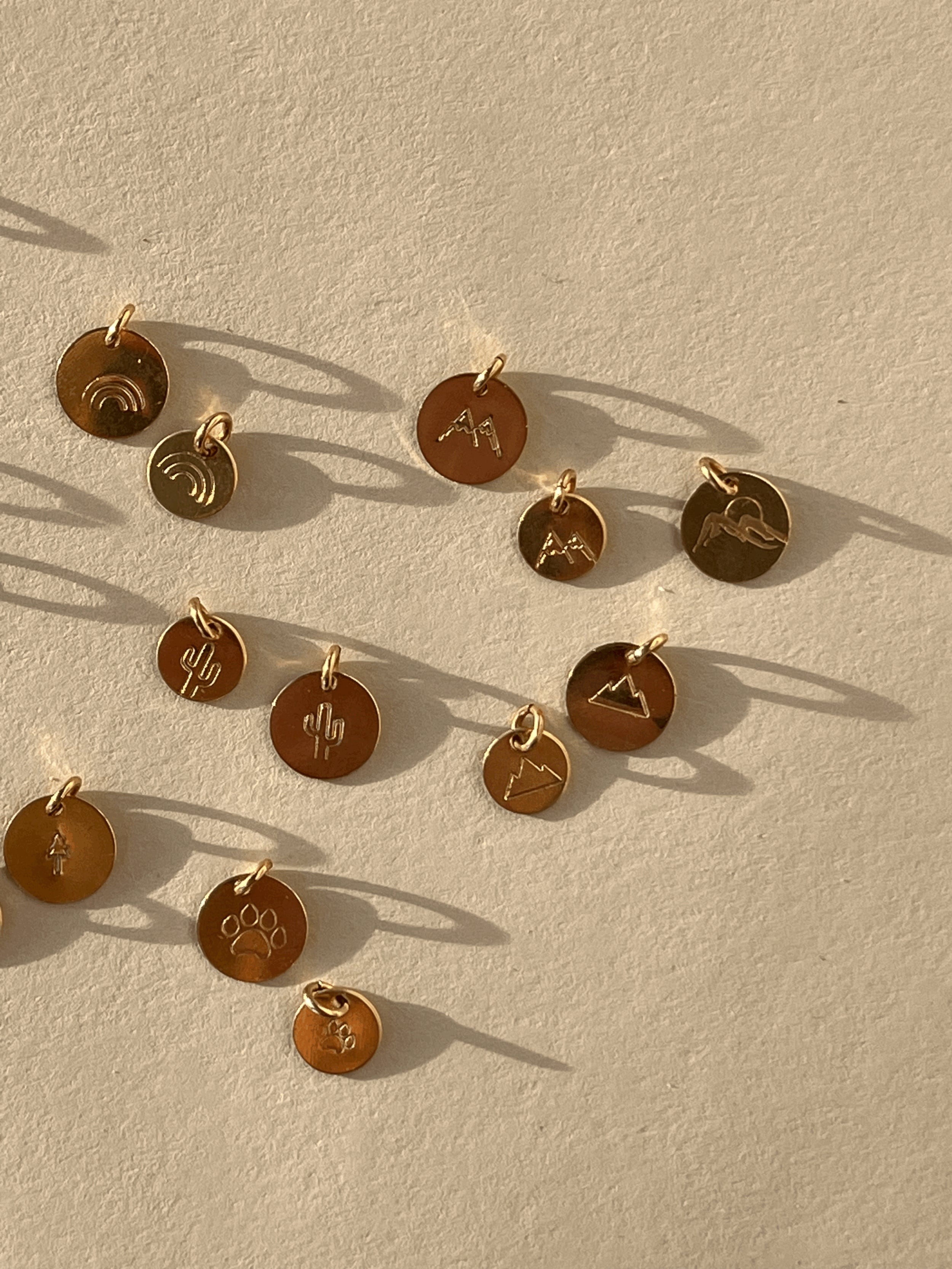 Gold-filled disc charms displayed on a rock, featuring tree, paw print, rainbow, cactus, three-peak mountains, two-peak mountains in 7mm and 9mm sizes, and mountains with sun design in 9mm only