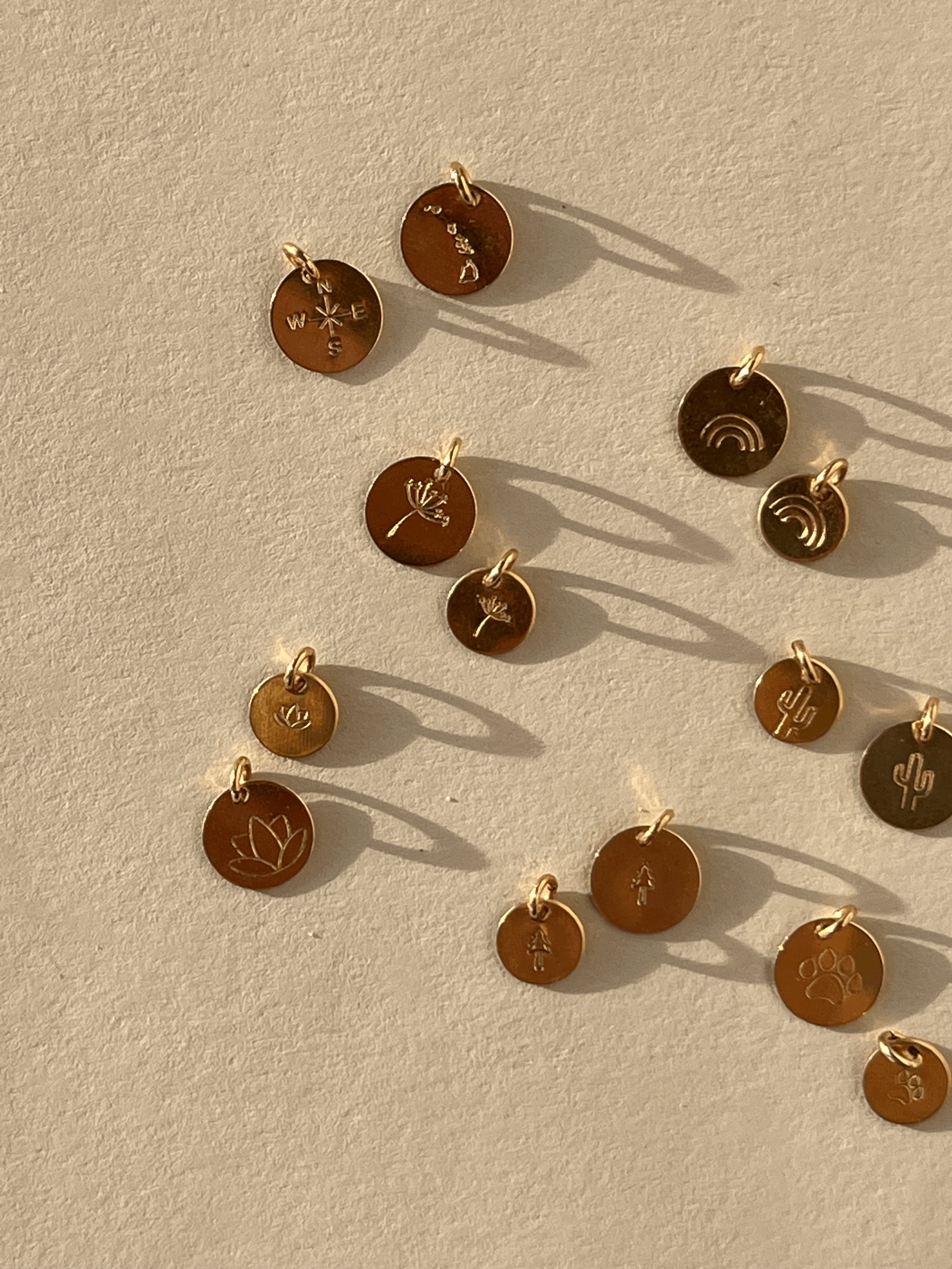 Gold-filled disc charms displayed on a rock, including tree, dandelion, lotus, paw print, and rainbow designs in 7mm and 9mm sizes, plus Hawaiian Islands and compass designs in 9mm only