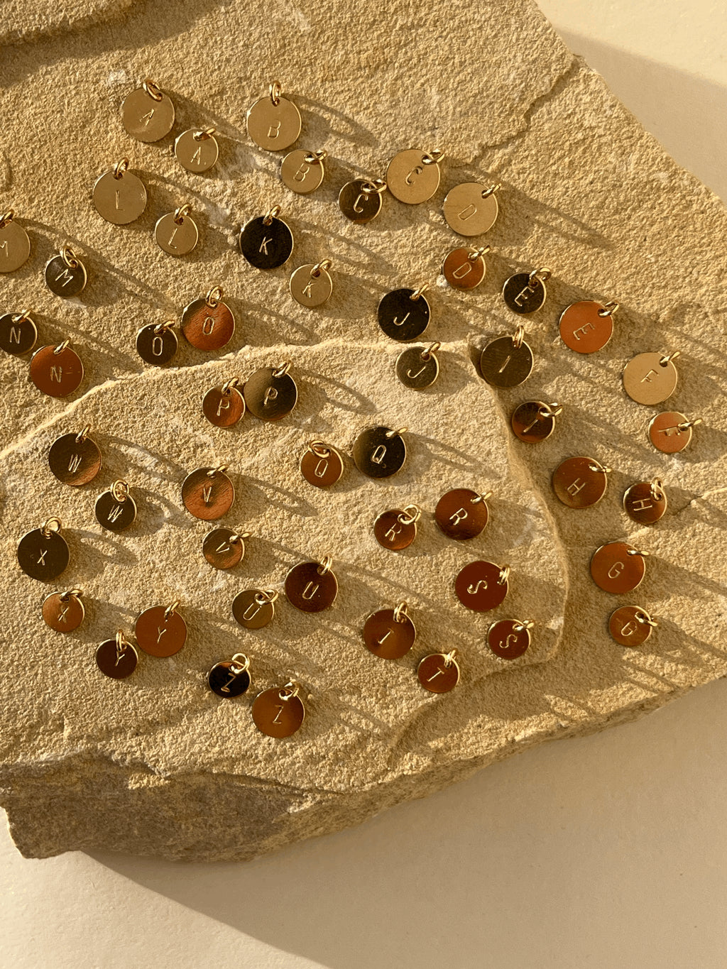 All twenty-six uppercase letters stamped on yellow gold-filled 7mm and 9mm disc charms, displayed without chains on a natural rock to show sizing and style