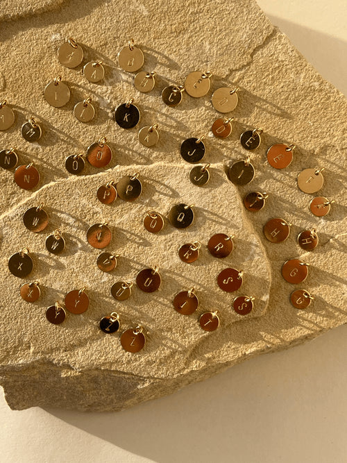 All twenty-six uppercase letters stamped on yellow gold-filled 7mm and 9mm disc charms, displayed without chains on a natural rock to show sizing and style