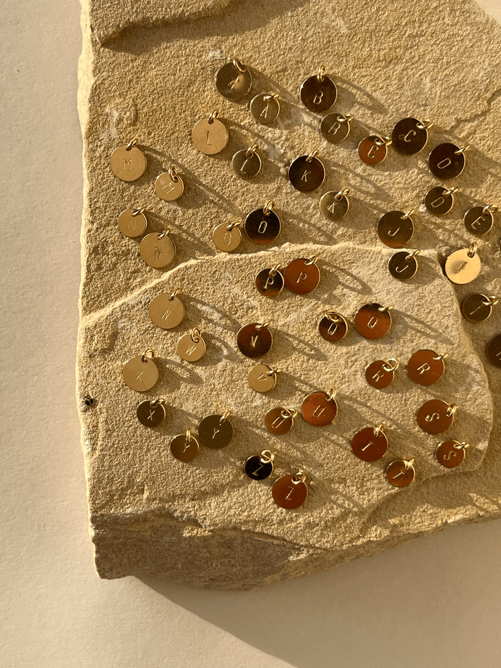 Different angle of all twenty-six uppercase letters stamped on yellow gold-filled 7mm and 9mm disc charms, displayed without chains on a natural rock to show sizing and style