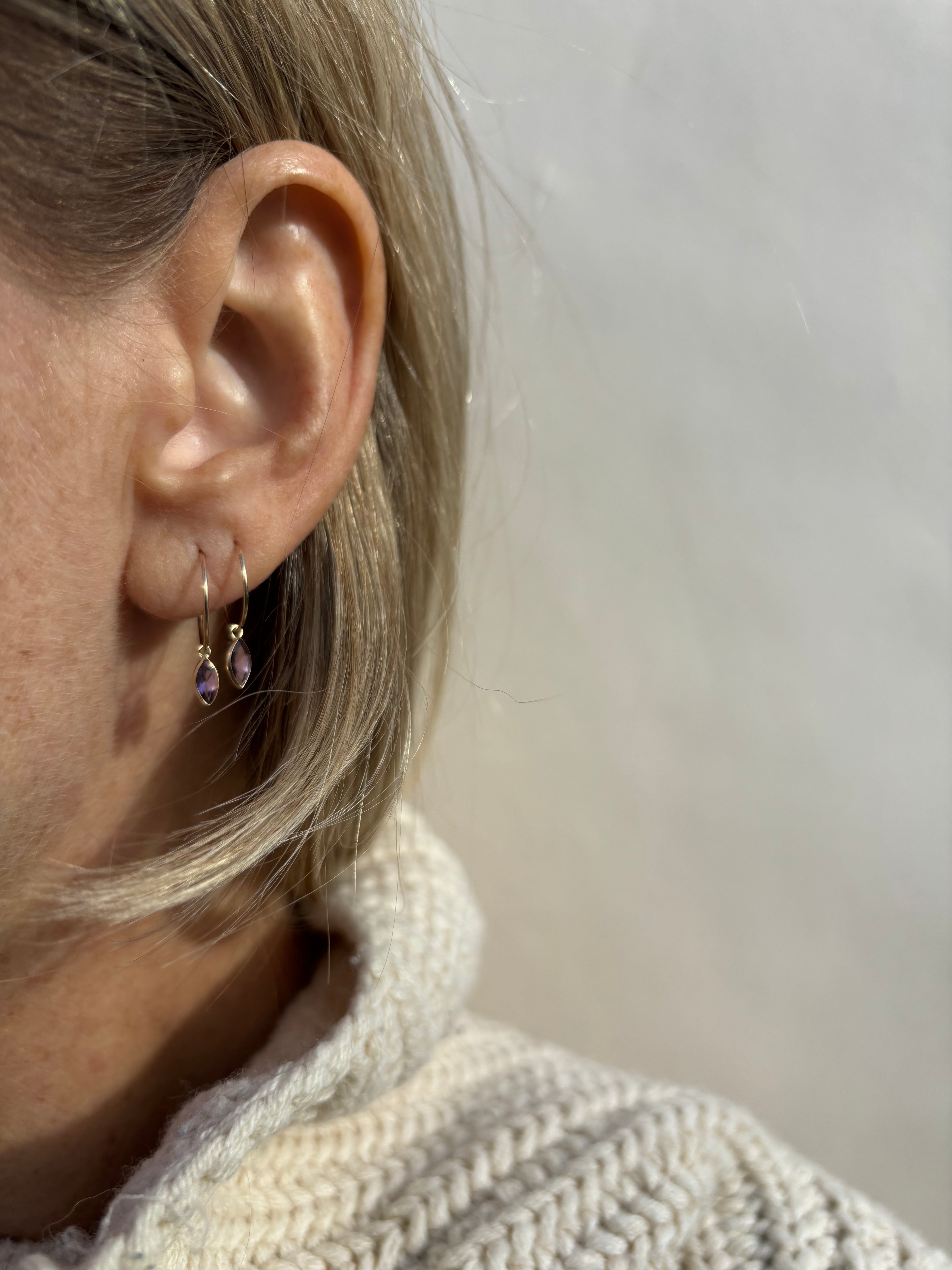 Close-up of model wearing Thalken 14k solid gold 12mm hoop earring with a small marquise-shaped natural amethyst drop in the first ear piercing, paired with a 10mm hoop with a matching marquise amethyst drop in the second piercing
