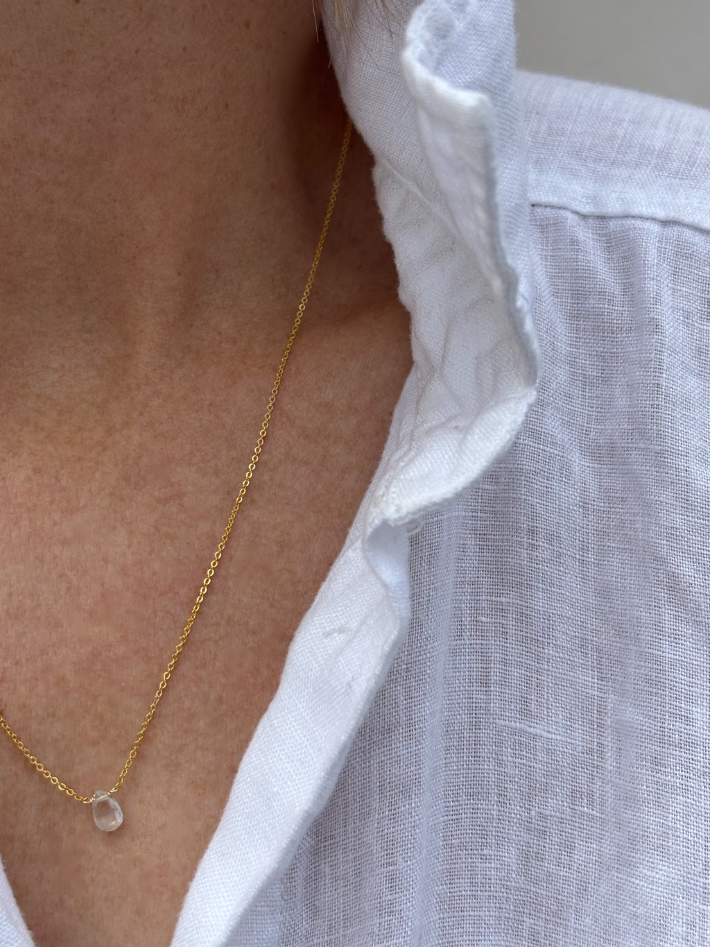 Close-up of dainty Thalken aquamarine drop necklace on woman wearing white linen button down shirt