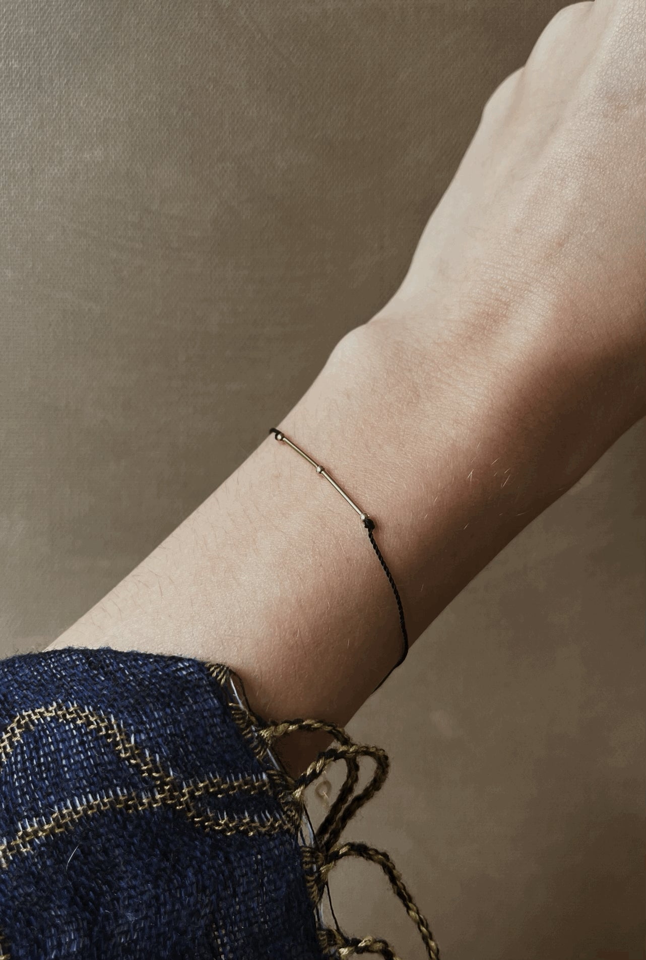 Thalken® Balance mantra bracelet with delicate black cord and gold-filled findings, shown on a slender wrist wearing an ornate blue shirt sleeve against a neutral backdrop
