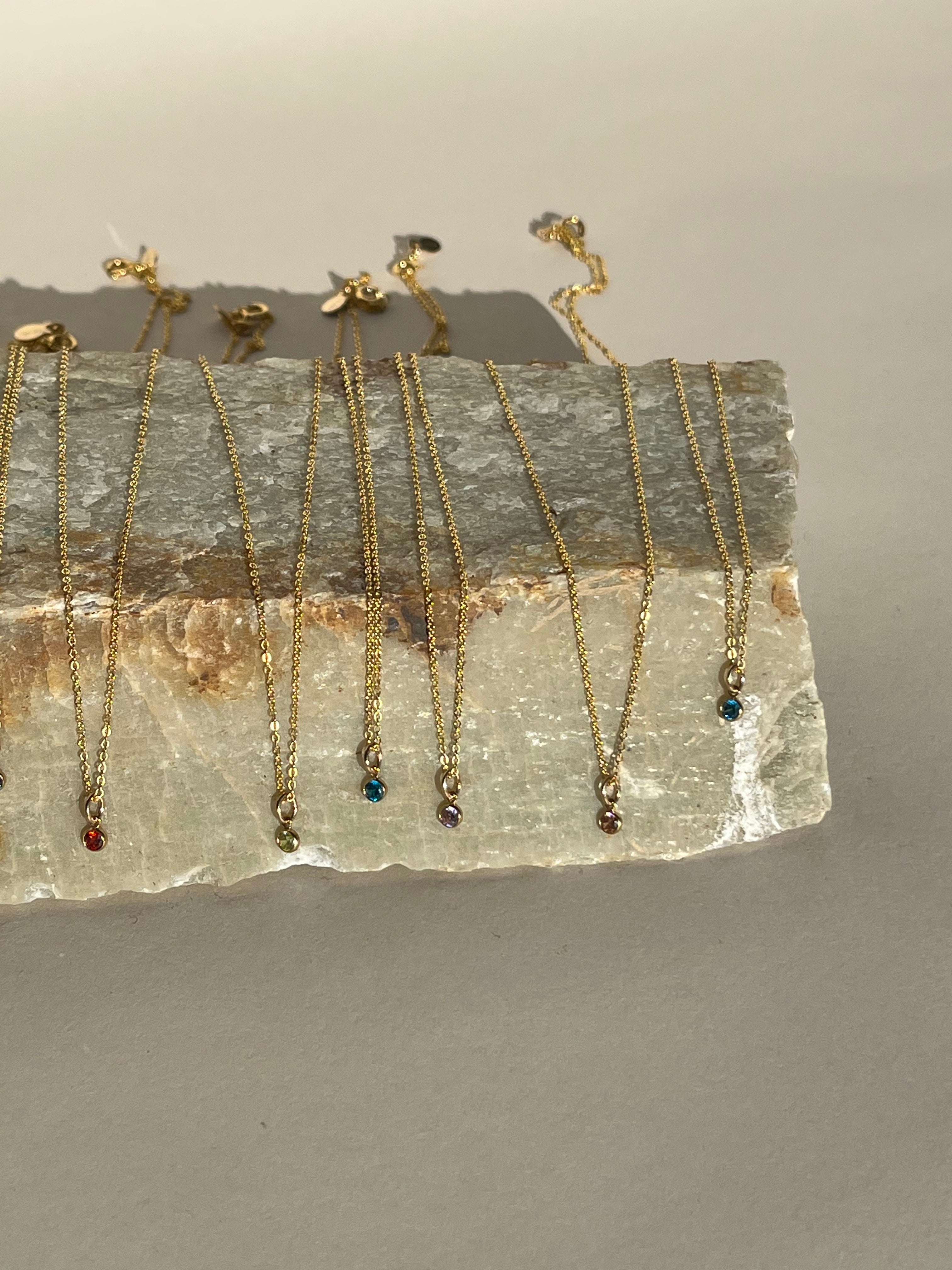 August through December dainty birthstone necklaces with 3mm gold-filled bezel-set cubic zirconia charms displayed on natural stone