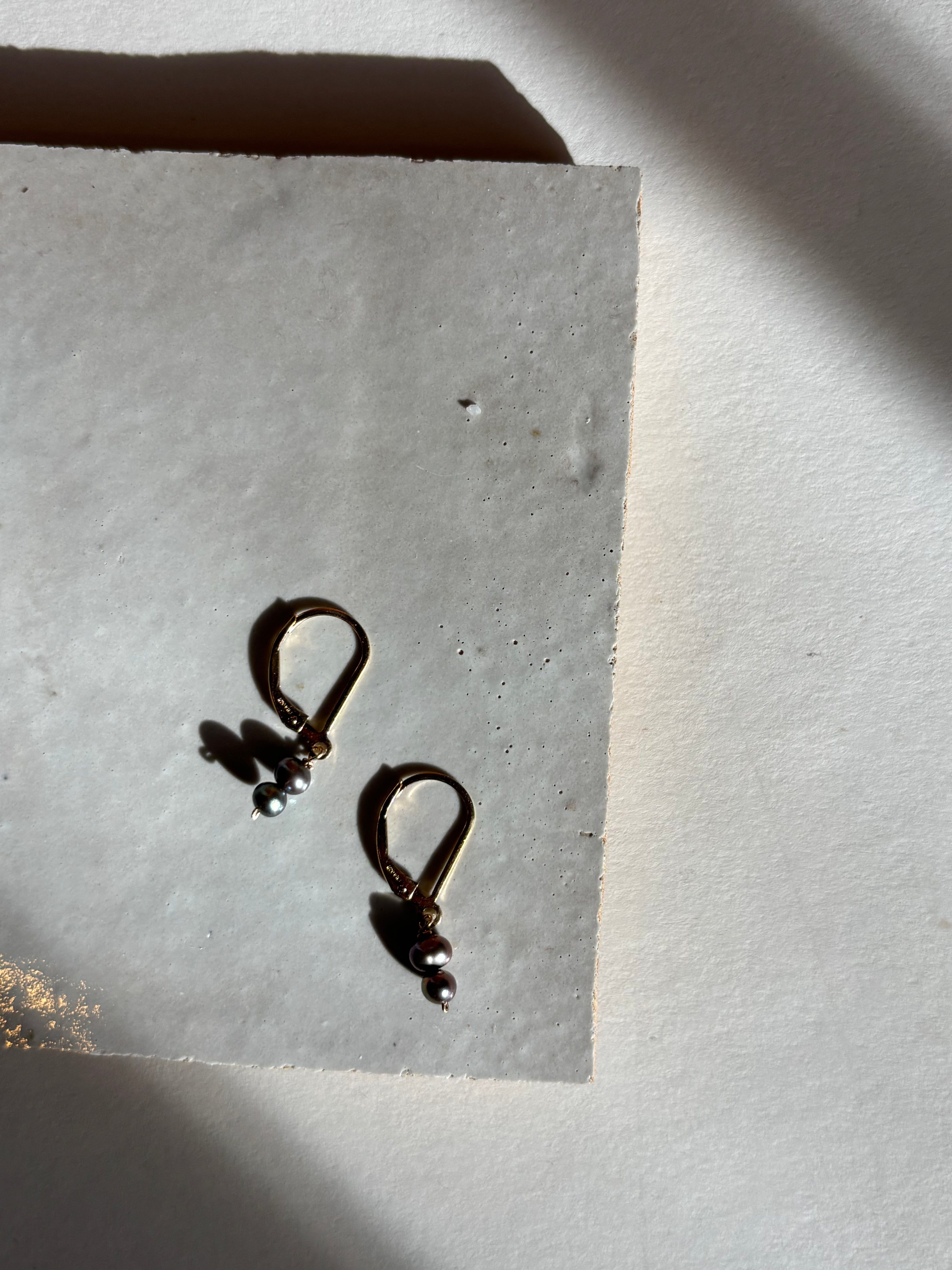 Delicate black double pearl drop Duet Extra Small earrings on an organic surface with warm, moody lighting showing the full earring design and pearl detail.