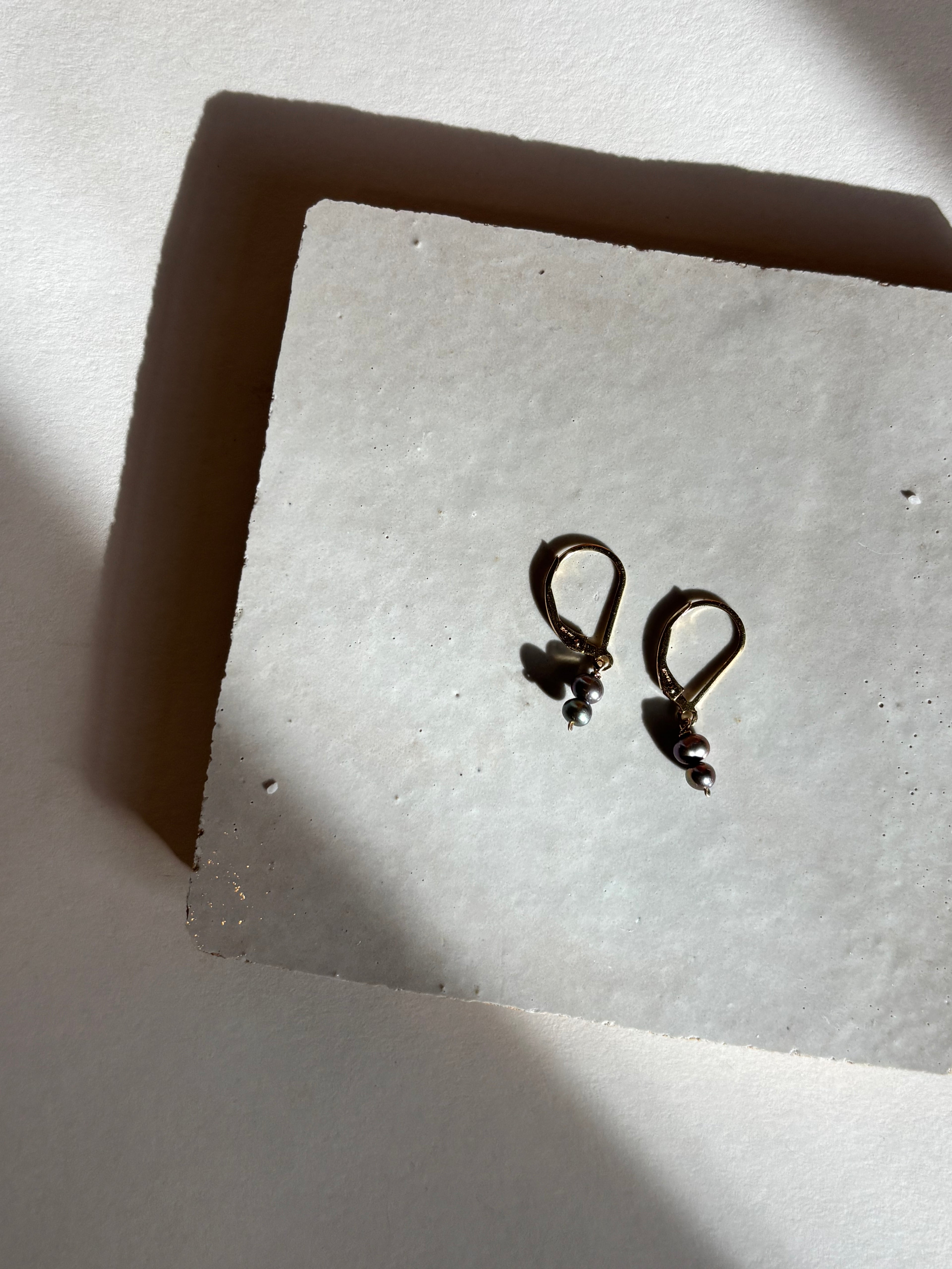 Delicate black double-pearl drop Duet Extra Small earrings on an organic tile.