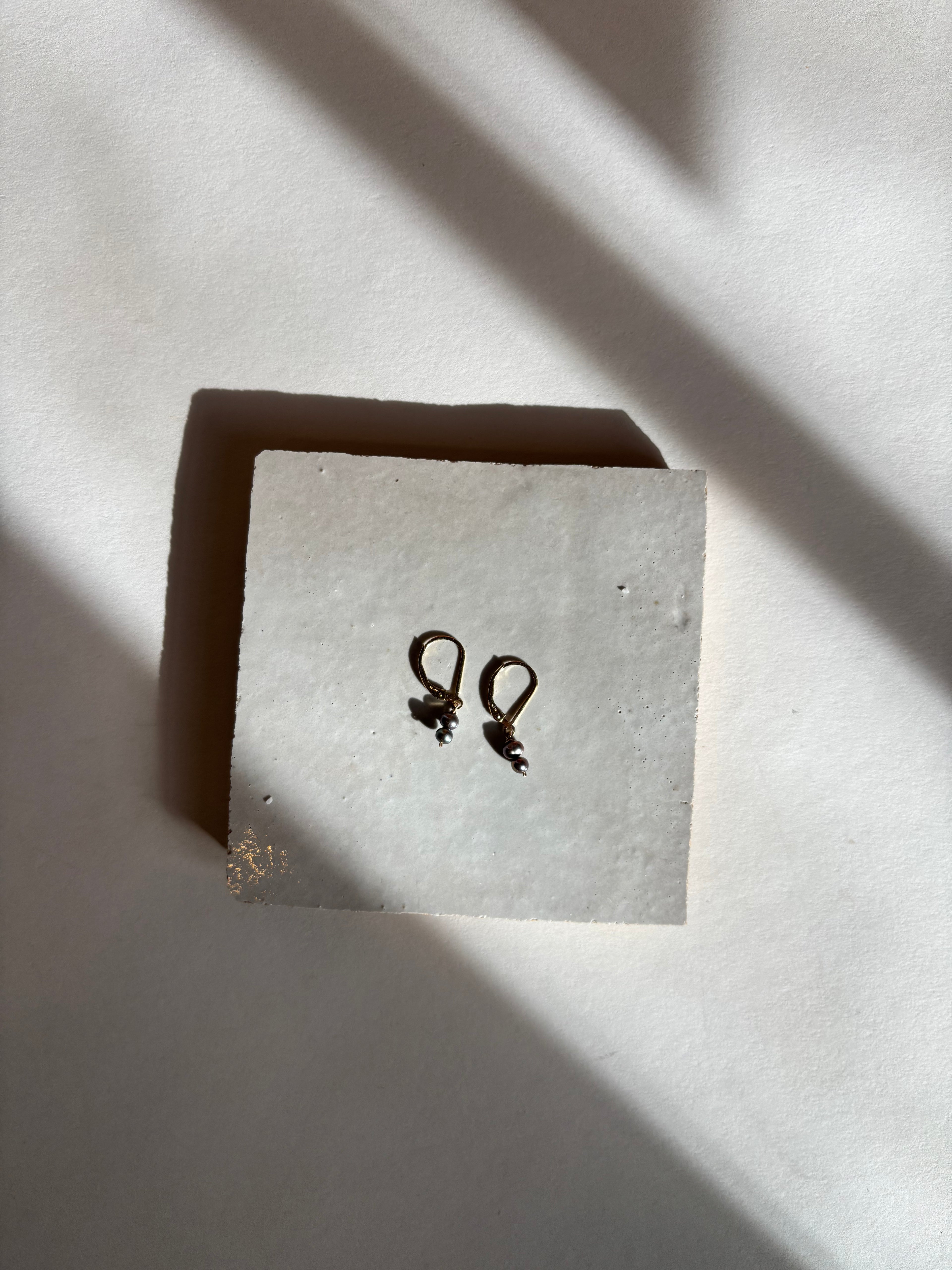 Delicate black double-pearl drop Duet Extra Small earrings on an organic surface with warm, moody lighting, showing the full earring design and pearl details. Bird’s-eye view shot.