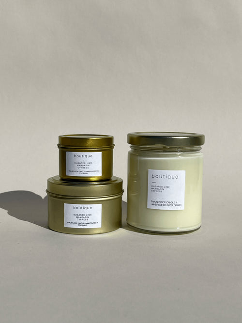 Photo of Thalken’s Boutique candle shown in three vessel options — sleek gold tins (2oz and 6oz) and a soft-glow glass jar (8oz). Bright and sunny scent profile with notes of sugared limes, mandarins, tropical fruits, and a touch of cypress