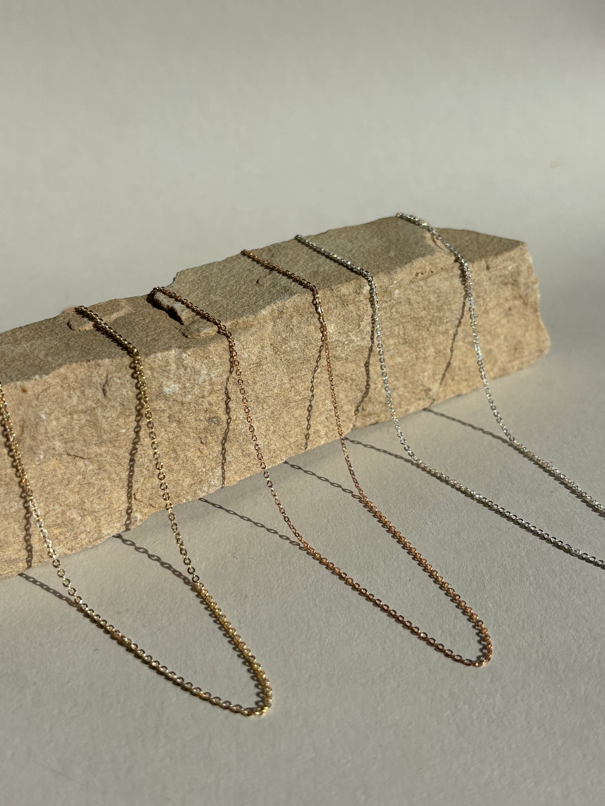 Three delicate chains—yellow gold-filled 1.3mm, rose gold-filled 1.1mm, and sterling silver 1.1mm—displayed together on a natural rock surface