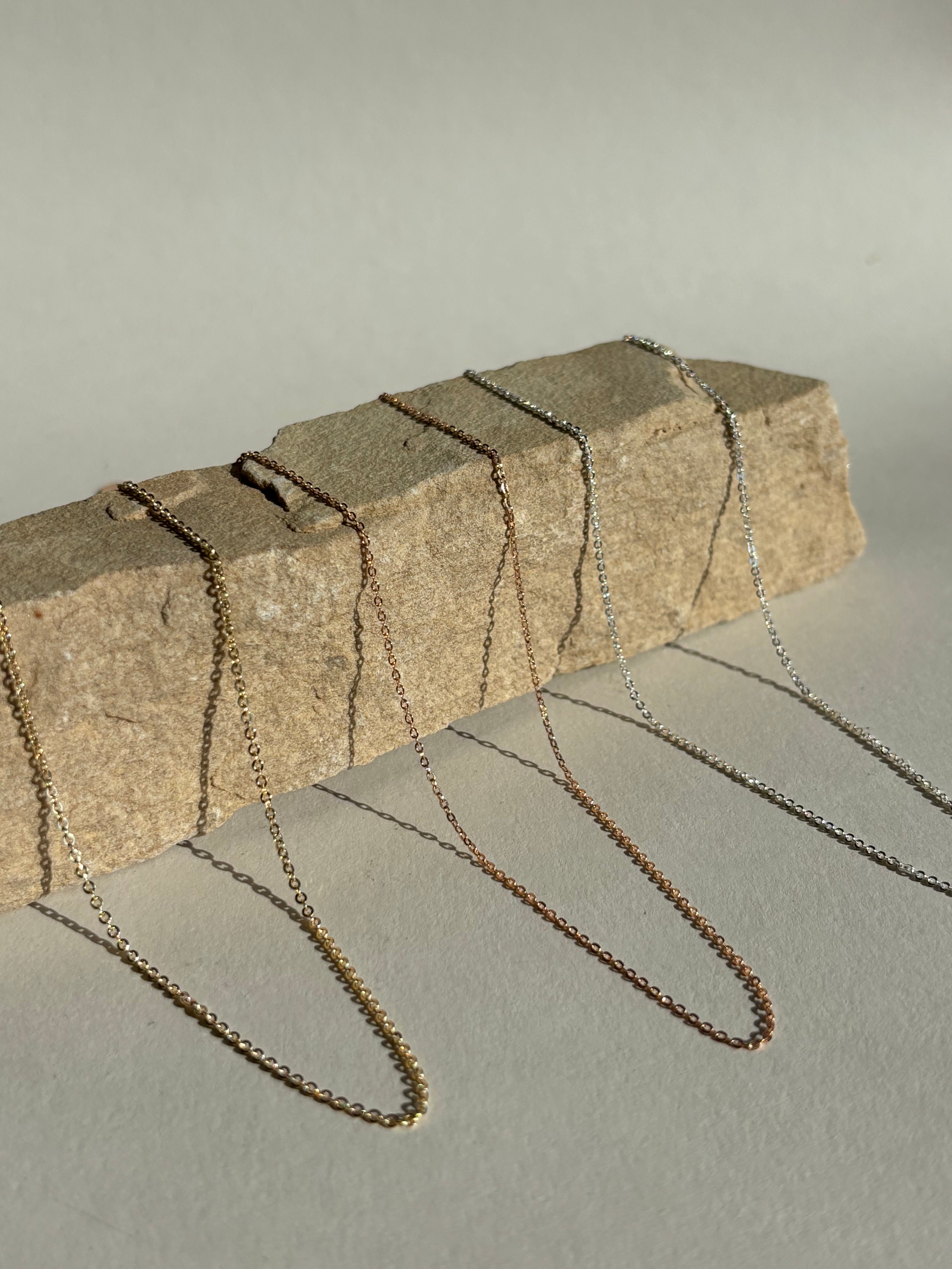 Three delicate chains—yellow gold-filled 1.3mm, rose gold-filled 1.1mm, and sterling silver 1.1mm—displayed together on a natural rock surface