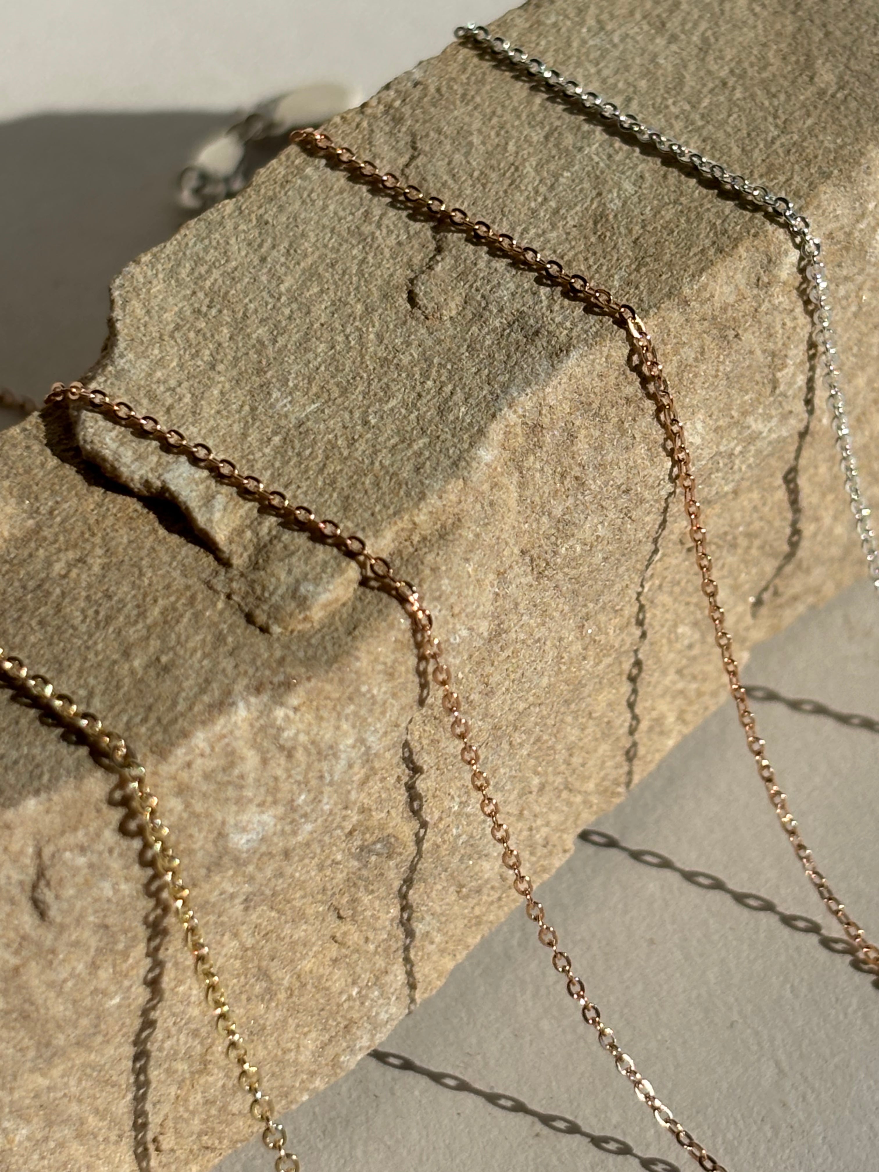 Detailed view of yellow gold-filled, rose gold-filled, and sterling silver delicate chains (1.3mm and 1.1mm) displayed side by side on a textured rock