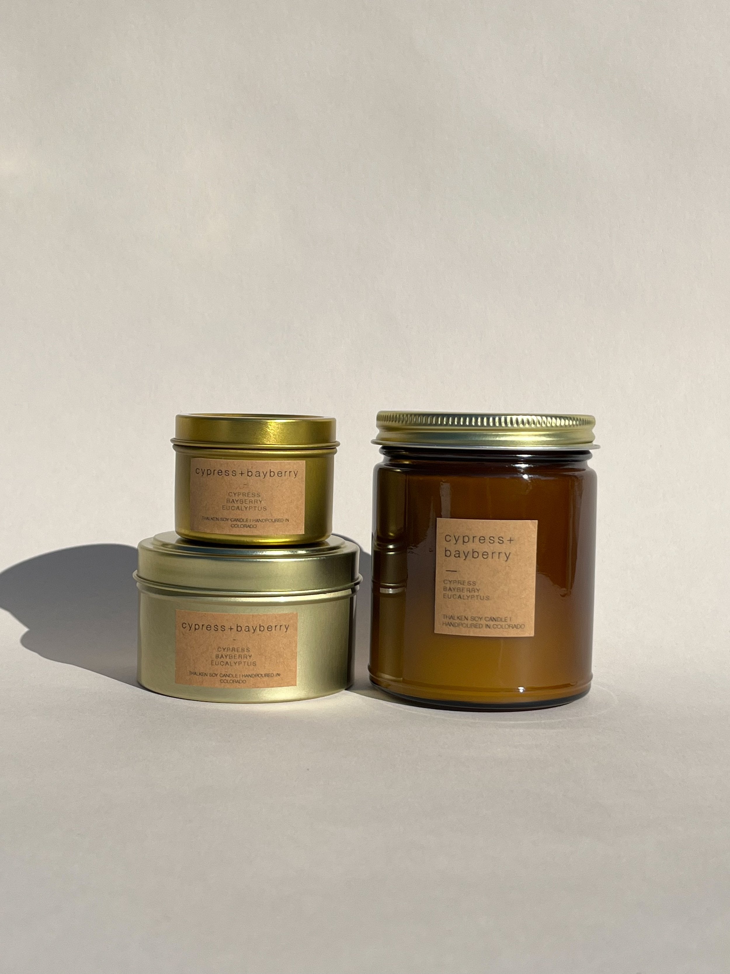 Thalken Cypress + Bayberry candles in three sizes — 2oz and 6oz gold tins and 8oz soft-glow glass jar. Scent profile: earthy, crisp cypress, bayberry, and eucalyptus, evoking the fresh scent of a pine forest