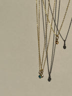 Blue, peach, and clear cubic zirconia birthstone charms on dainty gold-filled chains, suspended in mid-air against a natural background