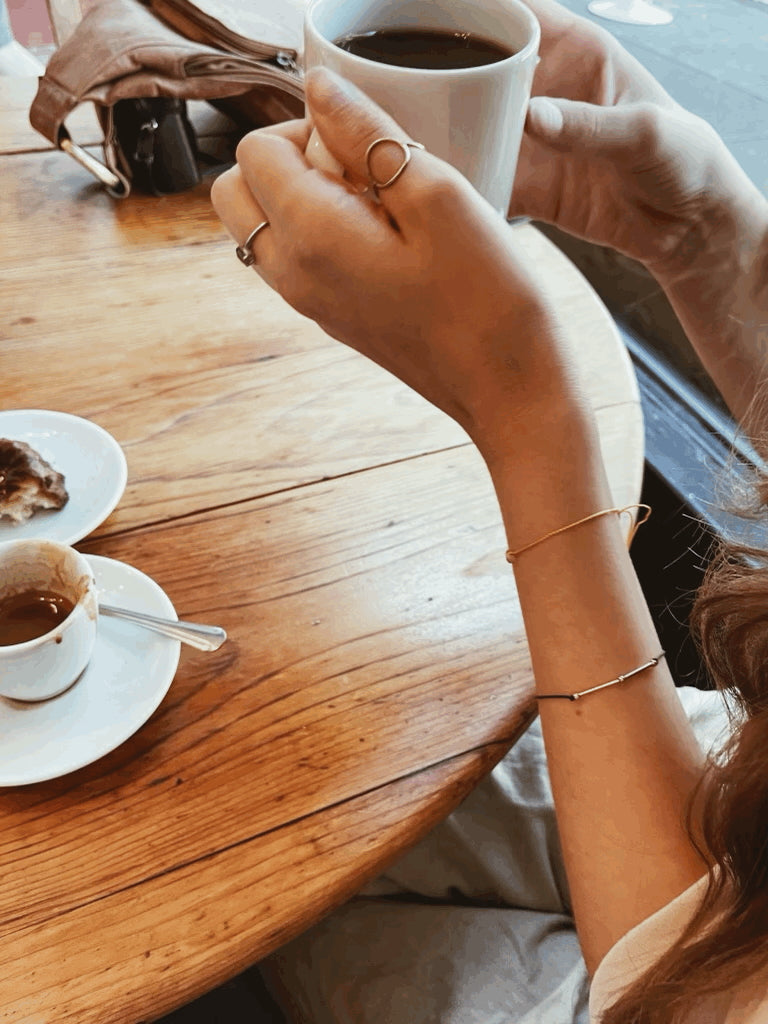 Drop of Sunshine Bracelet layered with the Balance Bracelet on model’s wrist, holding a cup of coffee inside a coffee shop