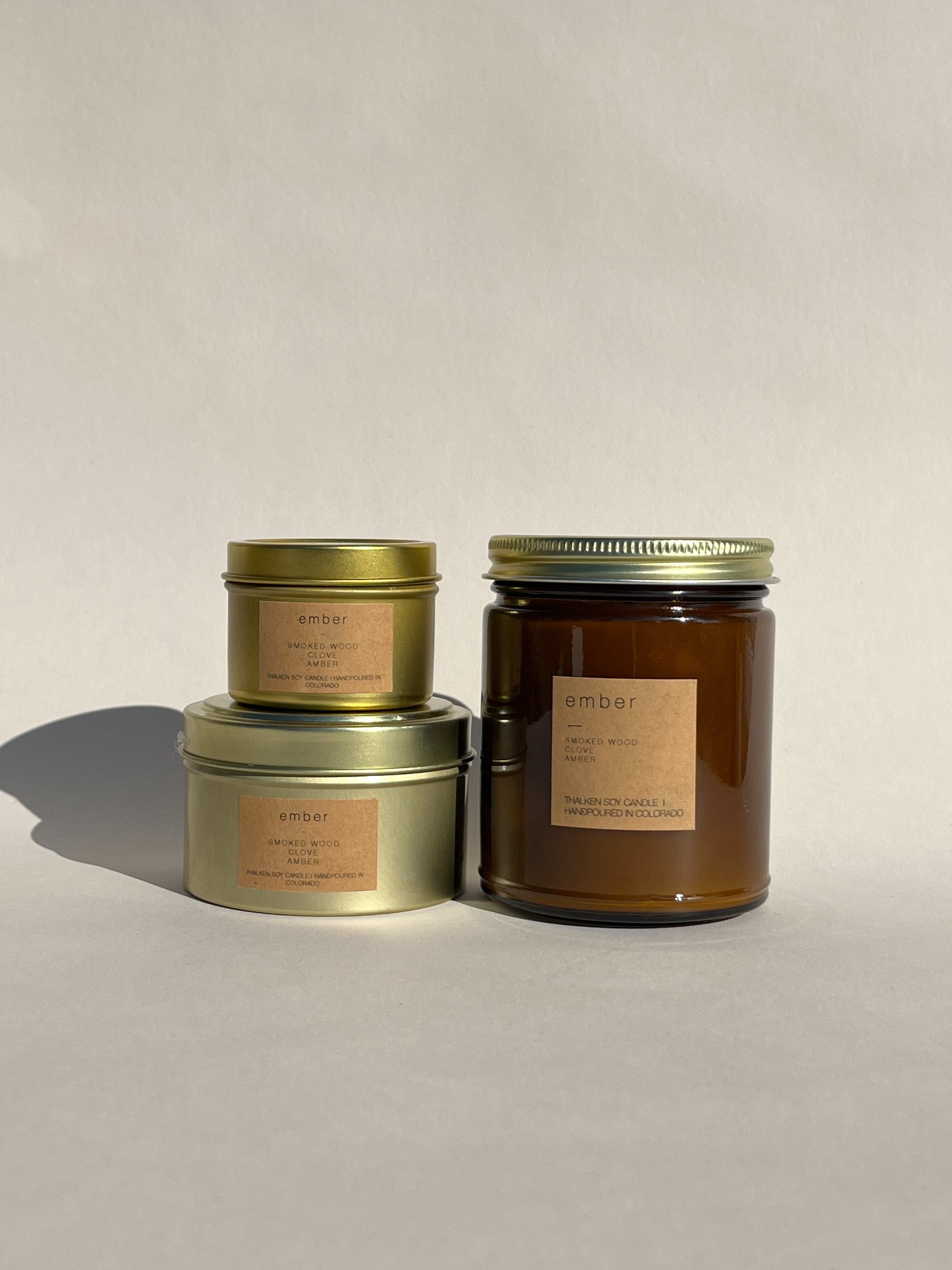 Thalken Ember candles in three sizes — 2oz and 6oz gold tins and 8oz soft-glow glass jar. Scent profile: smoked wood, clove, and amber.
