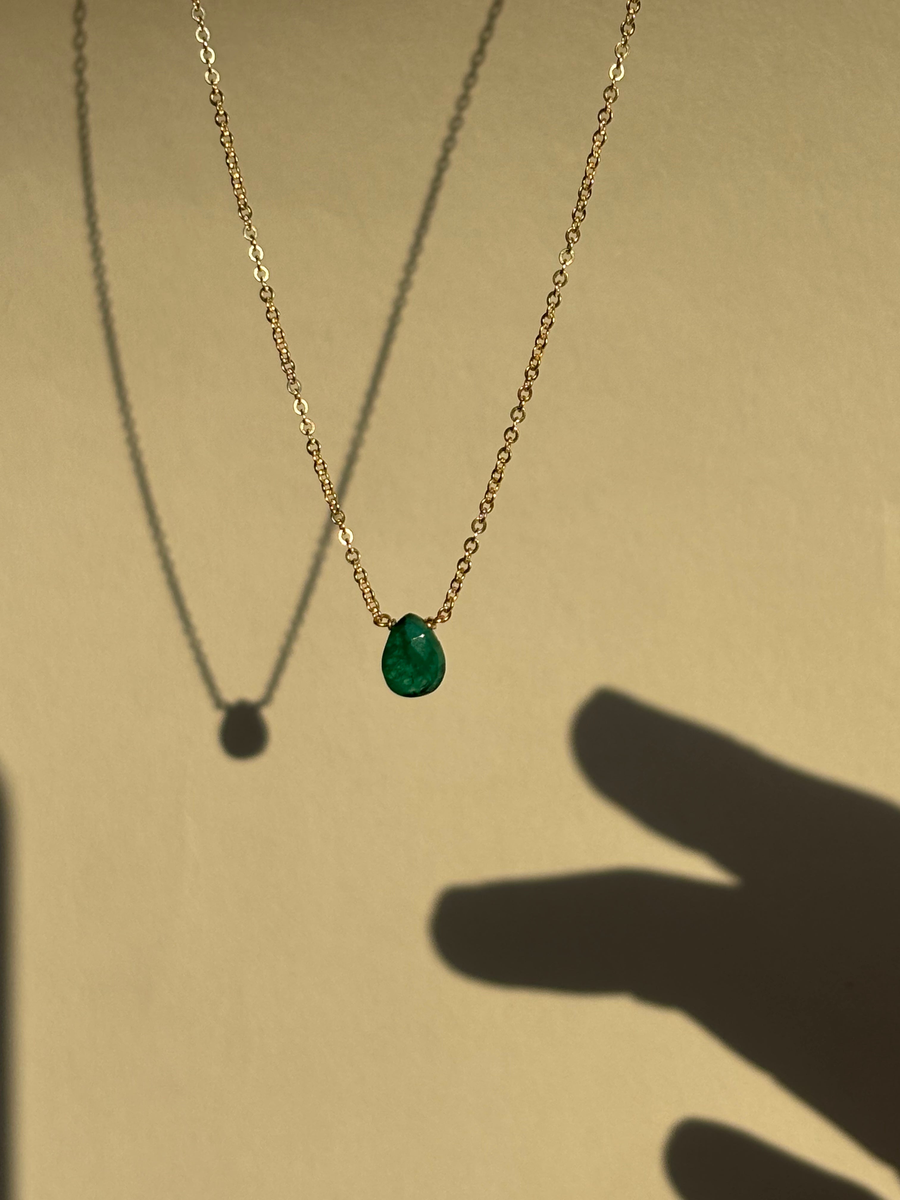 Thalken Emerald Drop Necklace with a green stone suspended in air, shadow of fingers reaching toward the necklace