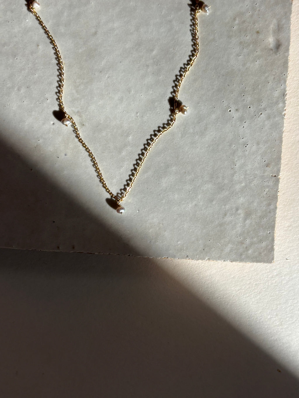 Close-up of the fog-toned freshwater pearl Whisper raindrop necklace on an organic tile surface with warm, moody lighting showing pearl spacing and fixed pearl details.