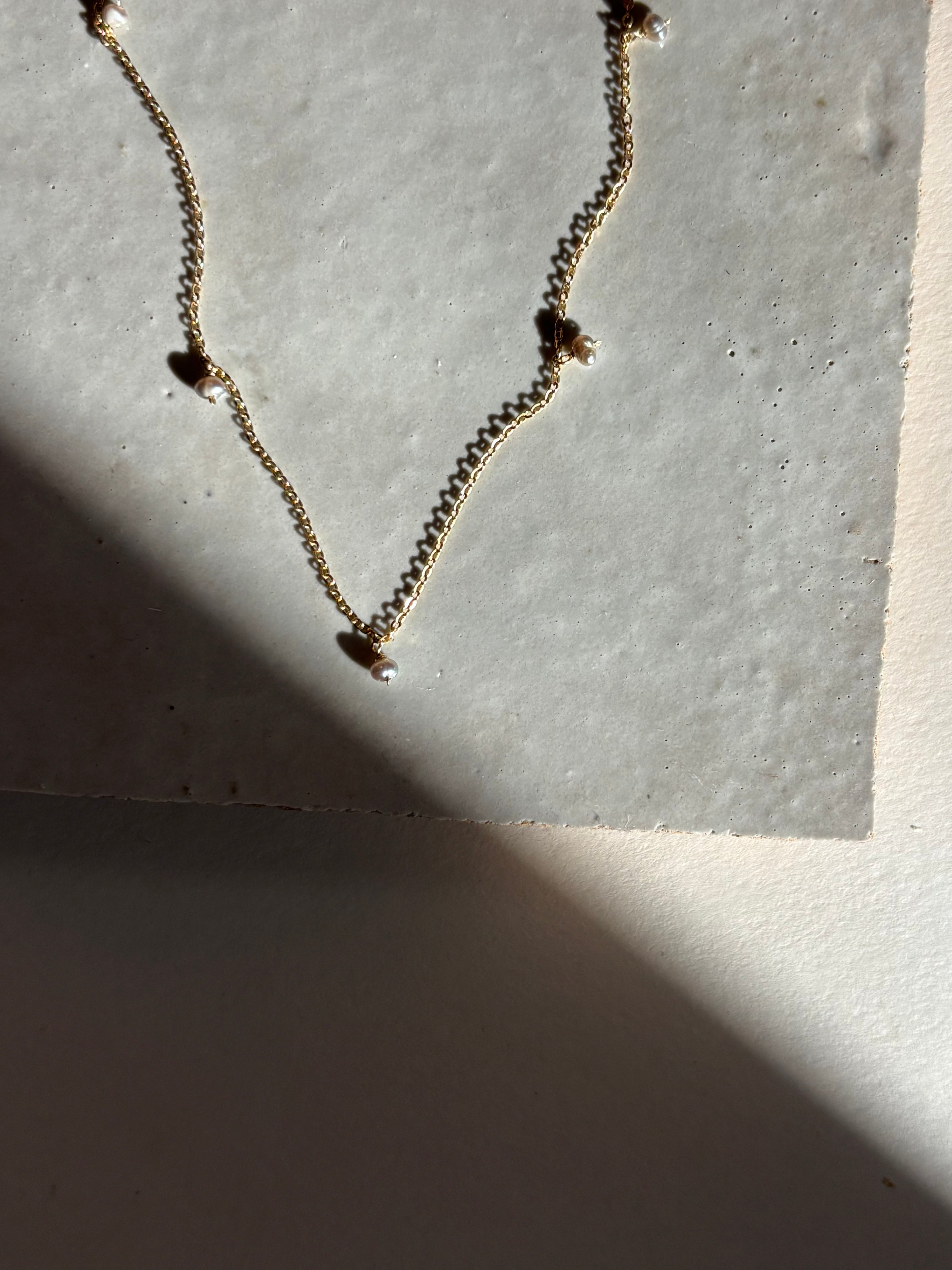 Close-up of the fog-toned freshwater pearl Whisper raindrop necklace on an organic tile surface with warm, moody lighting showing pearl spacing and fixed pearl details.