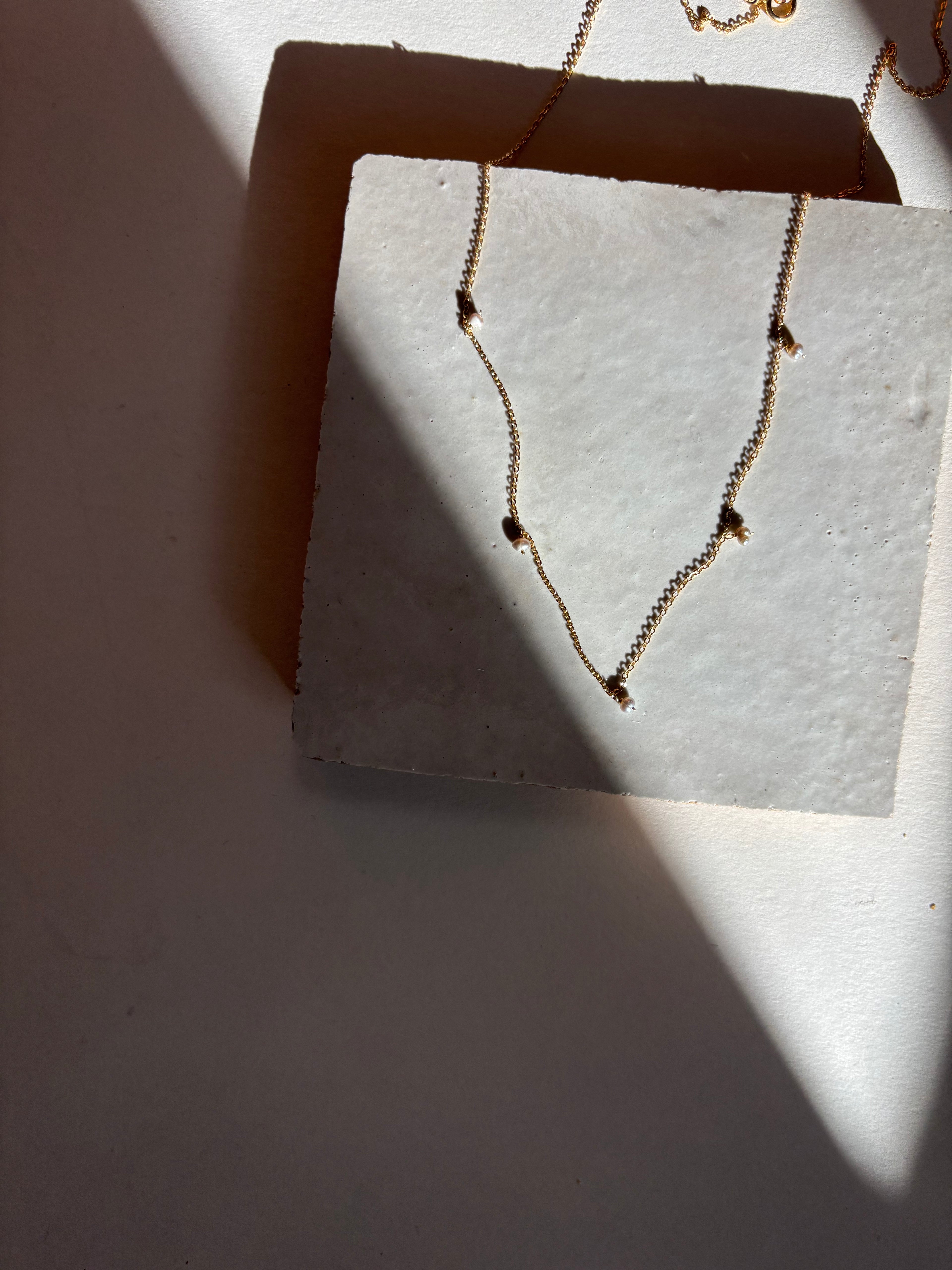 Fog-toned five pearl Whisper raindrop necklace displayed on an organic tile surface with warm, moody lighting showing the full necklace design and chain length.