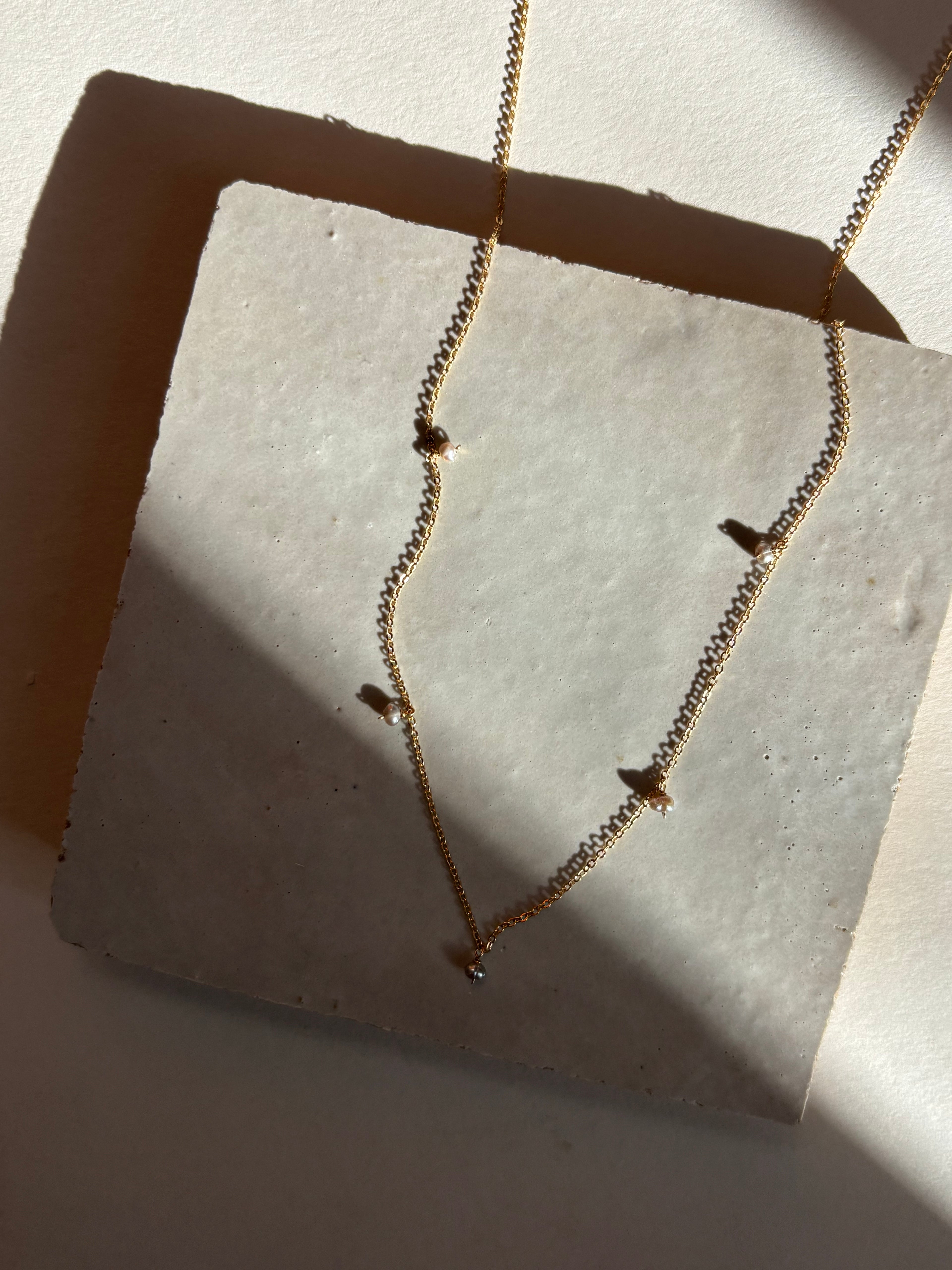 Fog-toned five pearl Whisper raindrop necklace displayed on an organic tile surface with warm, moody lighting showing the full necklace design and chain length.