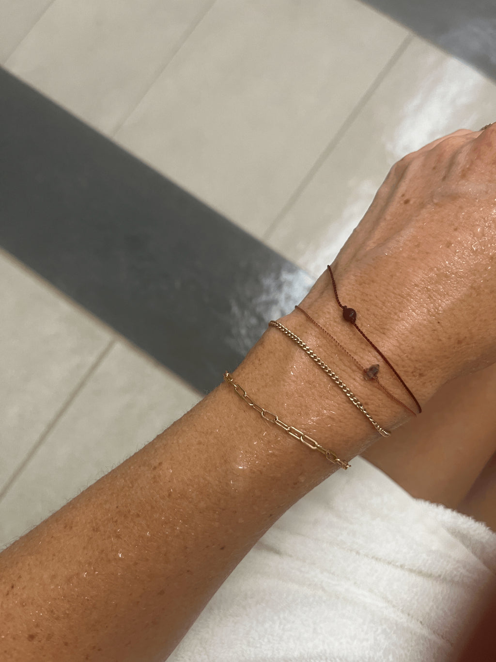 Tight bird’s-eye view of model in steam room with white towel, showing wrist stacked with the Thalken® Herkimer bracelet, Thalken® My Energy Is Sacred bracelet, Thalken® Original Small Link Paperclip Bracelet, and Thalken® Perfect Curb Chain Bracelet in yellow gold-filled