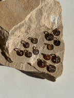 Rose gold-filled charms on rock featuring stamped letter H, tree, dandelion, lotus, paw print, rainbow, cactus, three-peak mountains, and two-peak mountains designs in 7mm and 9mm sizes in stamped letter T, Hawaiian Islands, compass, and mountains with sun designs