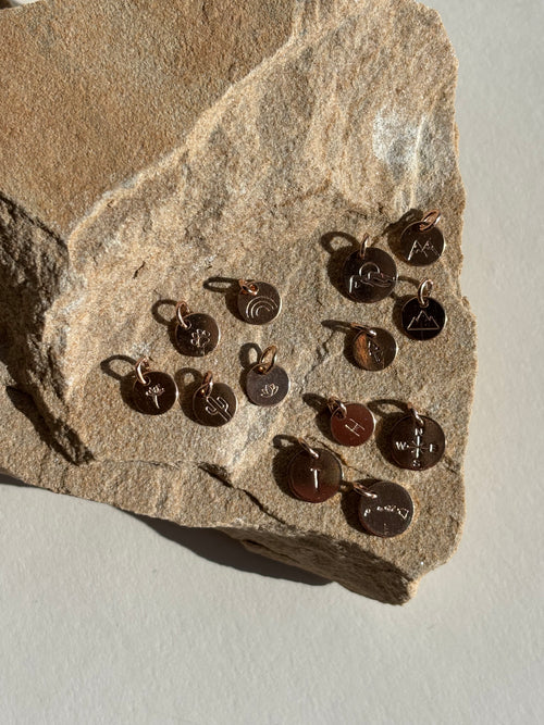 Rose gold-filled charms on rock featuring stamped letter H, tree, dandelion, lotus, paw print, rainbow, cactus, three-peak mountains, and two-peak mountains designs in 7mm and 9mm sizes in stamped letter T, Hawaiian Islands, compass, and mountains with sun designs